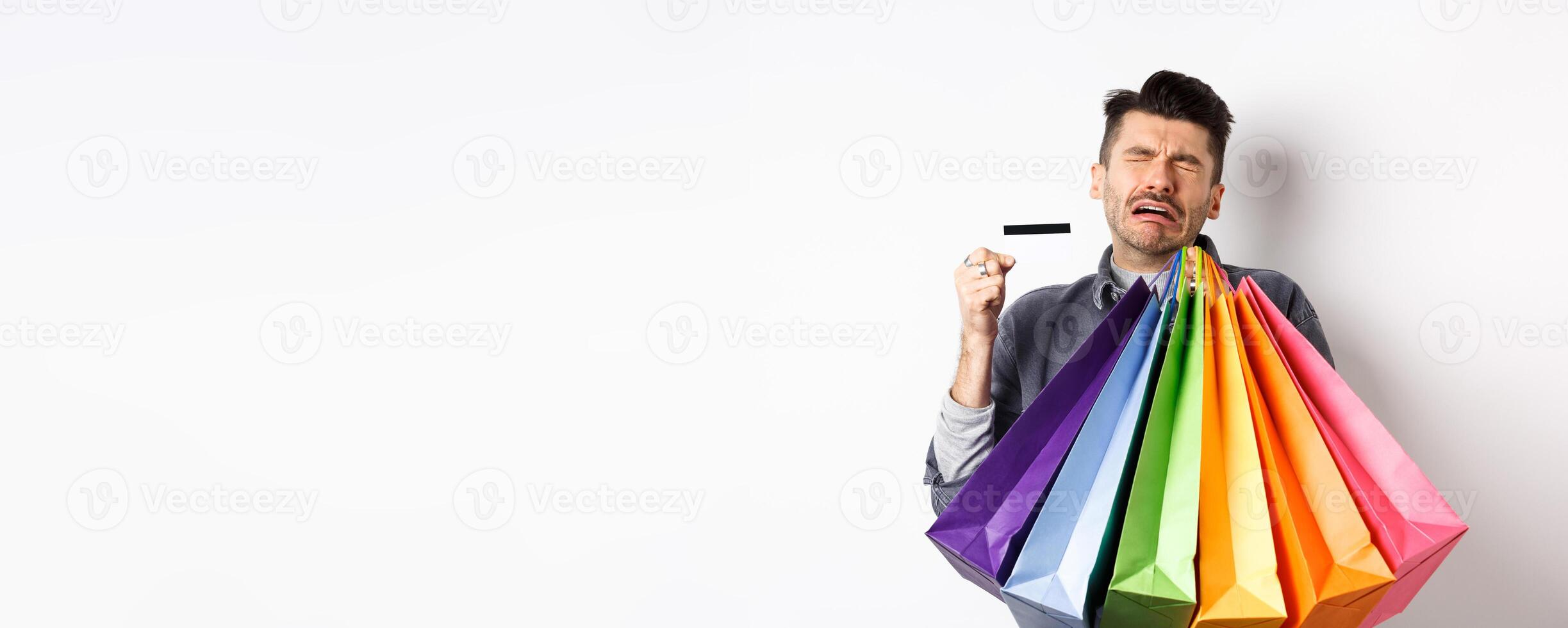 Sad crying man wasted all money on shopping, holding bags and showing empty credit card, standing against white background photo