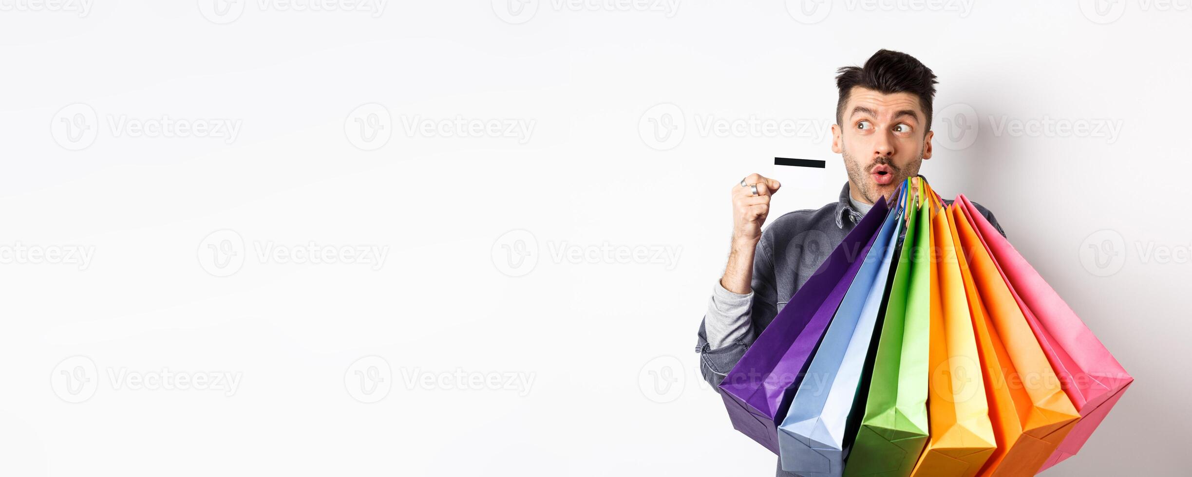 Excited young man buying in shop with plastic credit card, holding colorful shopping bags and looking aside at logo, standing against white background photo