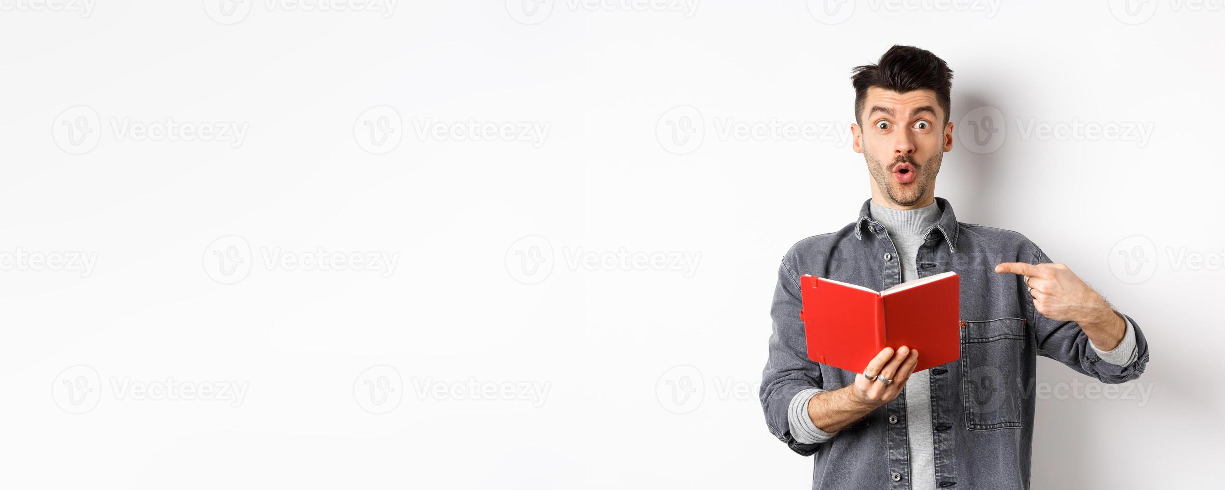 Excited guy read interesting thing in journal, pointing at planner and look impressed, standing against white background photo