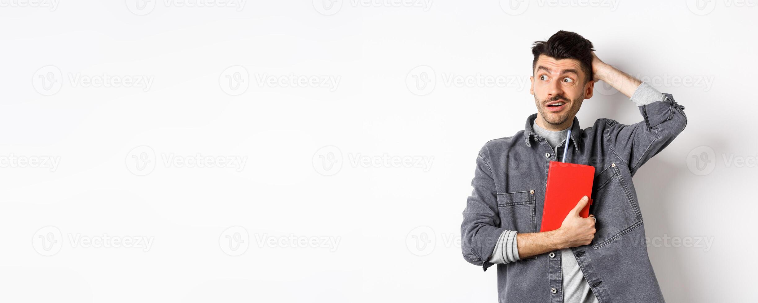 Confused guy scratch head and look aside at logo puzzled, holding red diary or planner, standing against white background photo