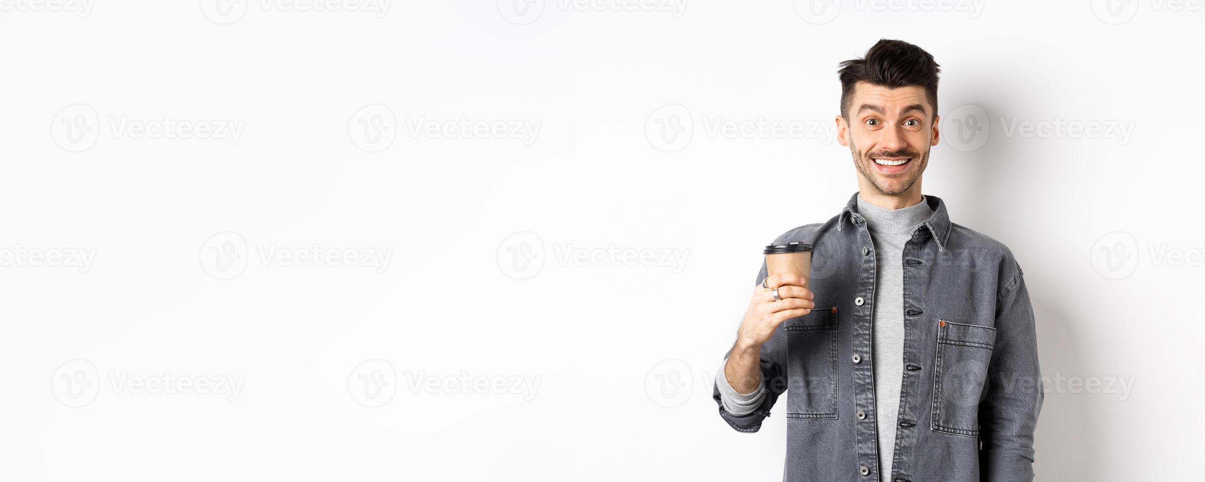 Enthusiastic handsome man holding coffee cup and smiling, drinking good drink from cafe takeout, standing against white background photo