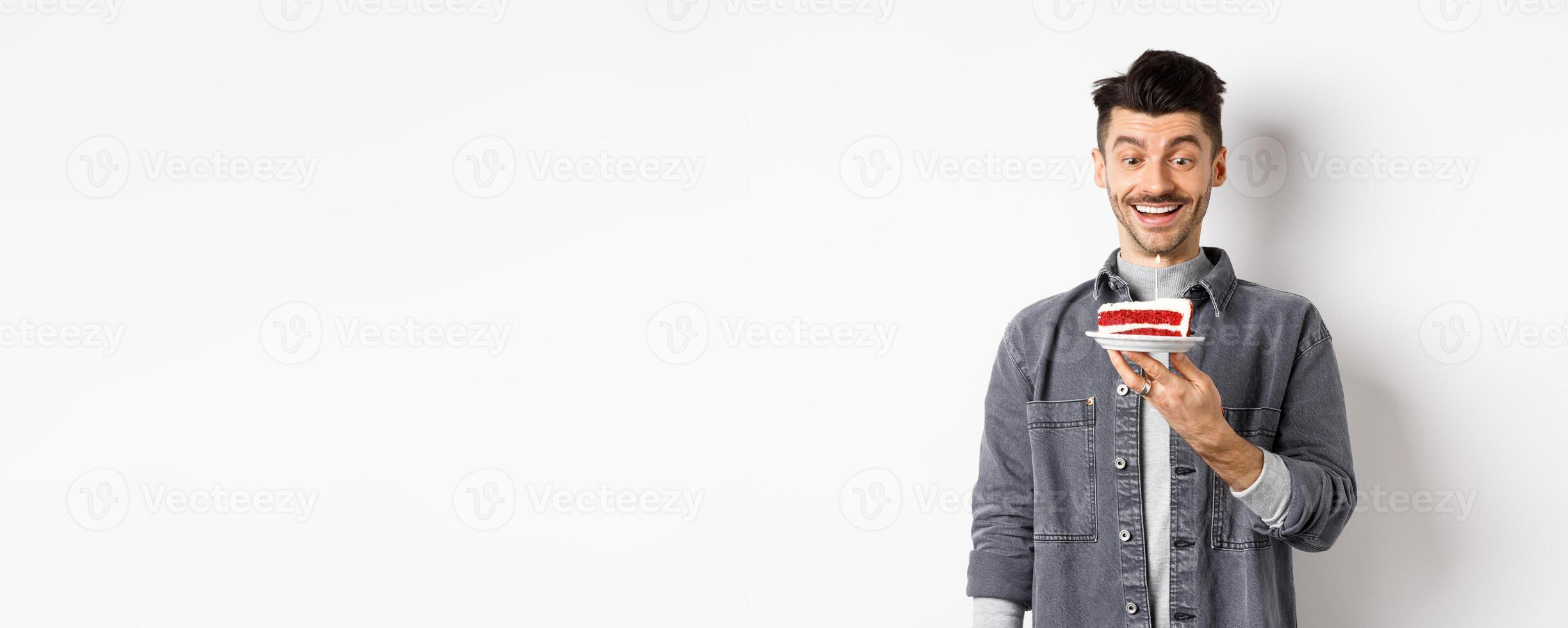 Happy birthday guy making wish on b-day cake, smiling and looking at lit candle excited, standing on white background photo