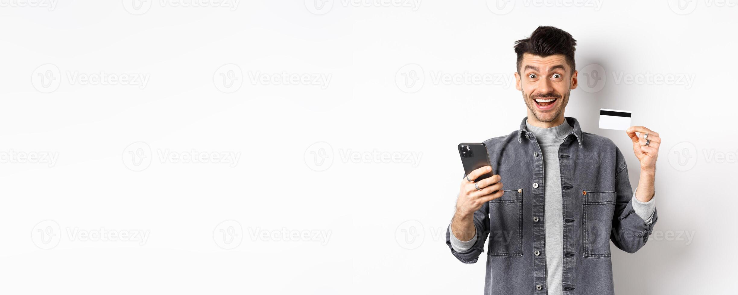 Online shopping concept. Excited man making purchase in internet, easy buying with plastic credit card and smartphone, smiling happy at camera, white background photo