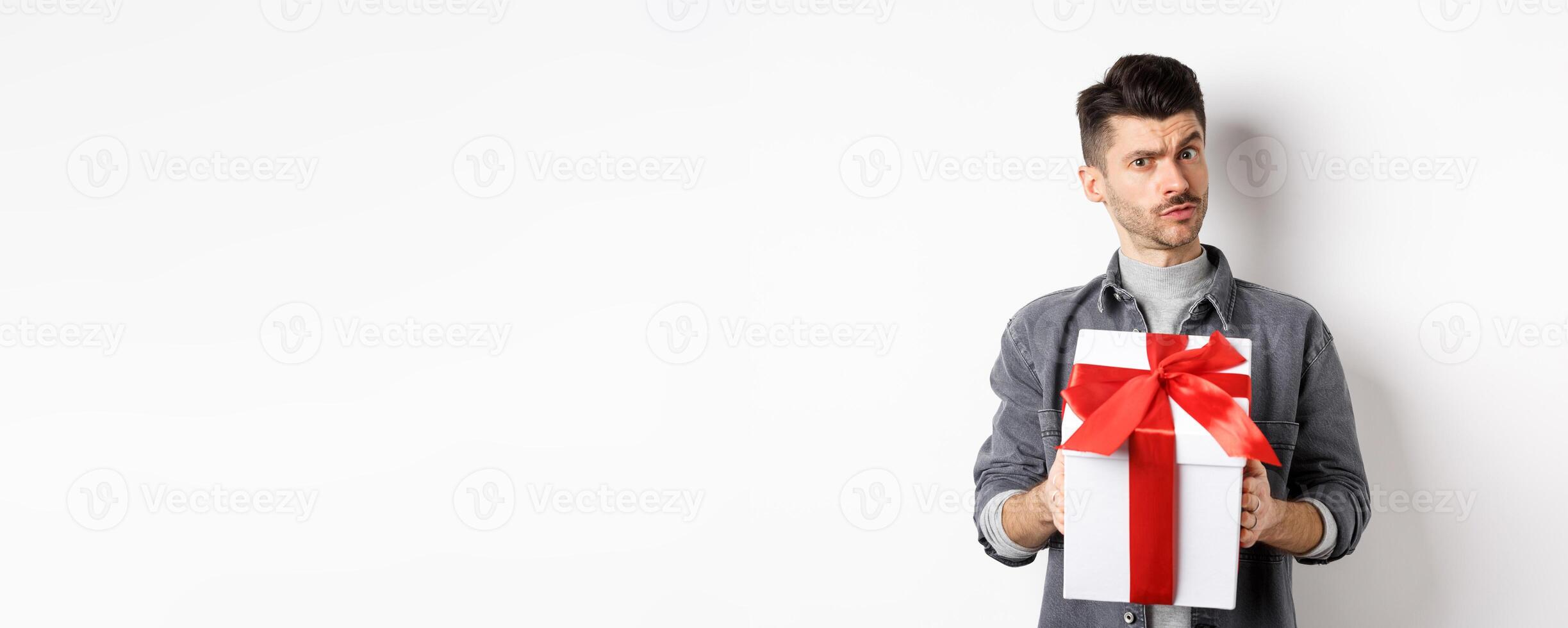 Surprised young man look with disbelief and hold surprise gift, raising eyebrow doubtful, being suspicious, standing on white background. Valentines day concept photo