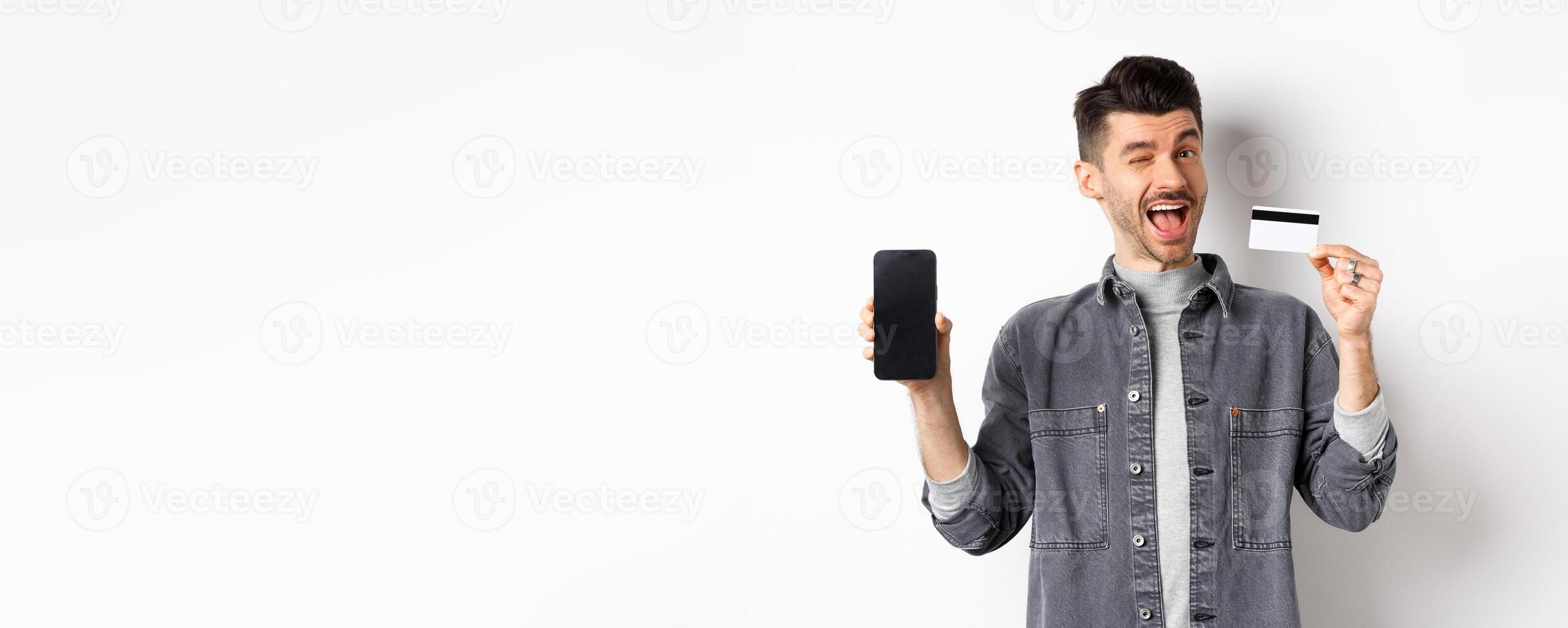 Cheerful handsome guy showing good deal, empty smartphone screen and plastic credit card, winking at you and smiling, recommending offer, white background photo
