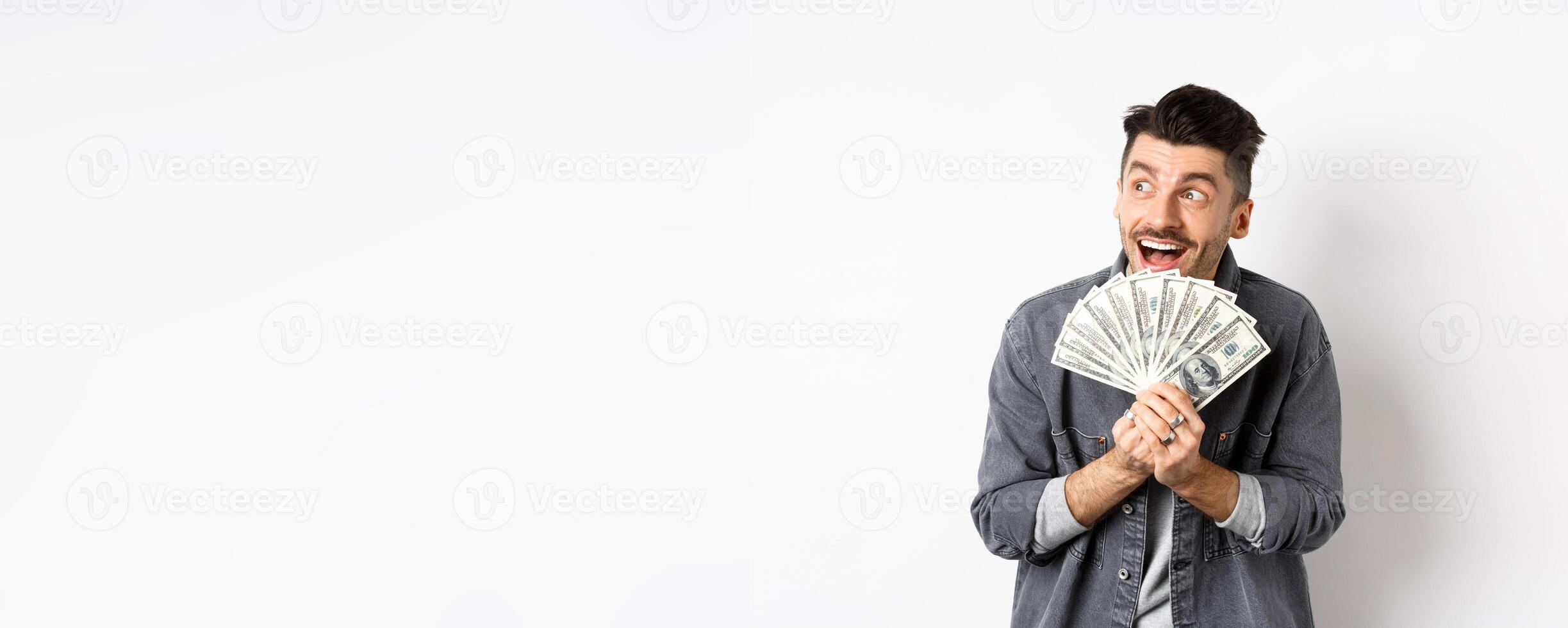 Rich happy man dreaming of shopping wish dollar bills in hands, looking aside pensive and holding money, standing against white background photo