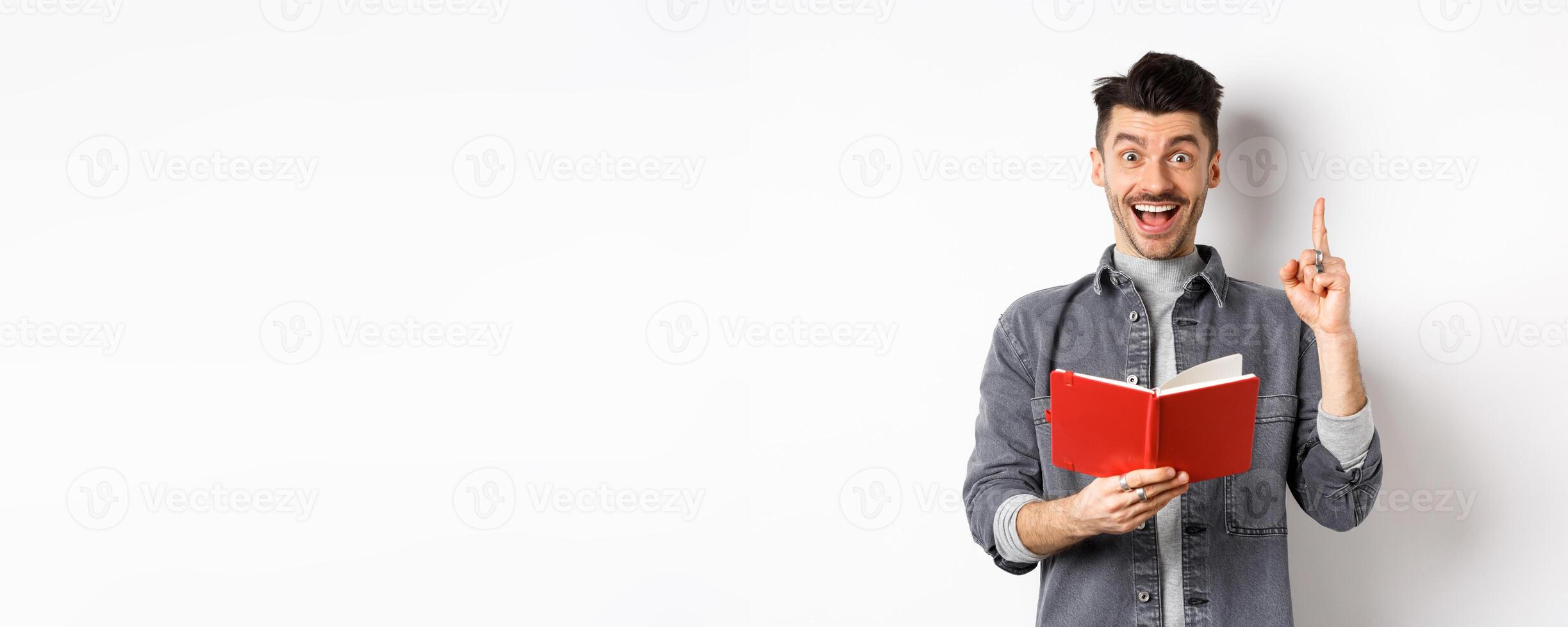 Excited guy pitching an idea while doing homework, raising finger and holding planner, standing against white background photo