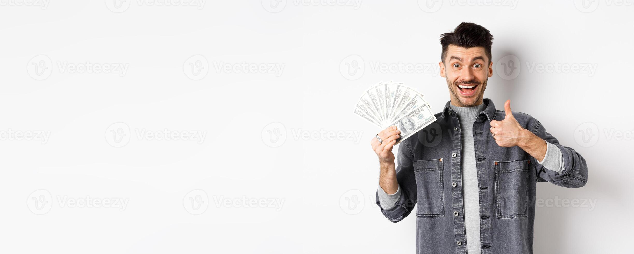 Image of excited man holding dollar bills and show thumbs-up with happy face, making money, standing on white background photo