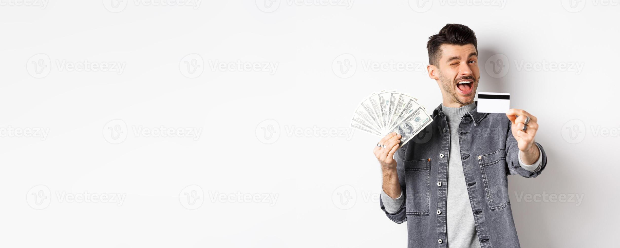 Image of handsome guy holding dollar bills but suggesting use plastic credit card, smiling friendly at camera, standing on white background photo