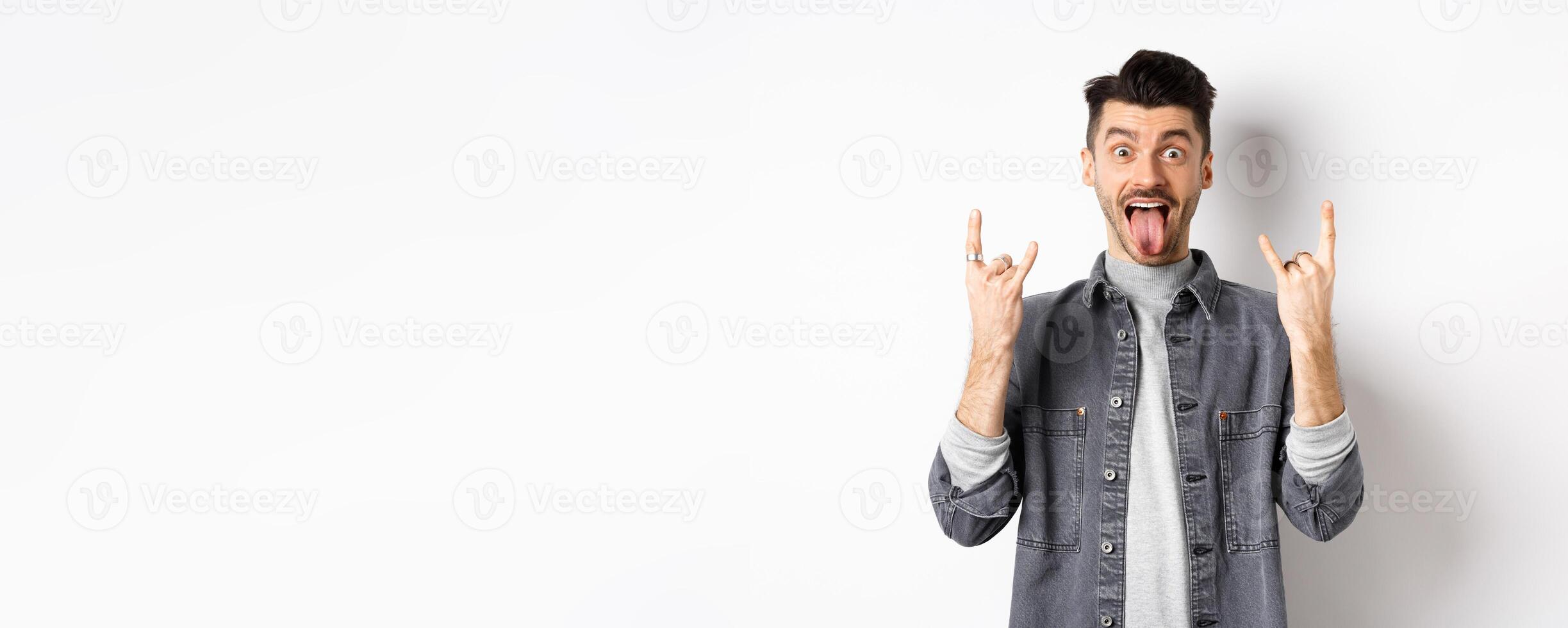 Excited funny guy showing tongue and heavy metal horns sign, enjoying party or event, having fun, standing on white background photo