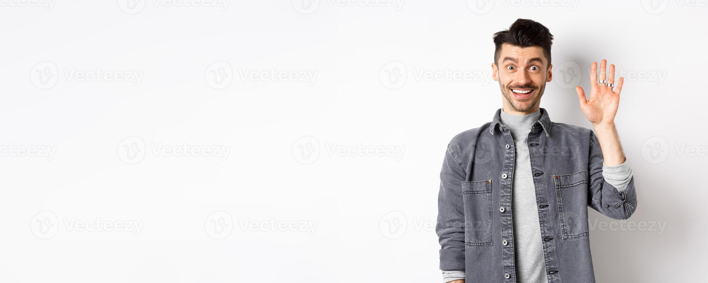 Friendly stylish man saying hello and waiving hand, smiling cheerful, greeting you with hi gesture, standing on white background photo