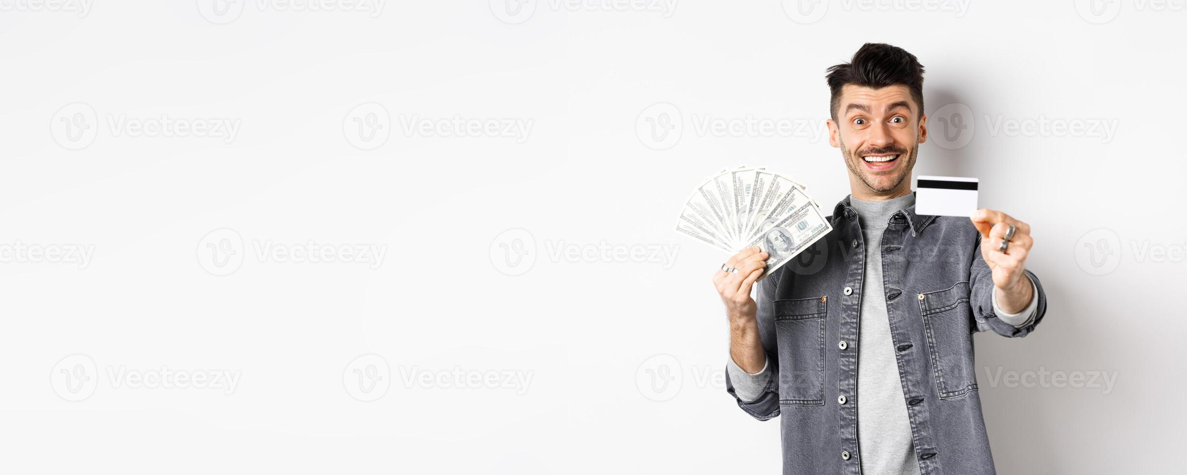 Image of handsome guy holding dollar bills but suggesting use plastic credit card, smiling friendly at camera, standing on white background photo