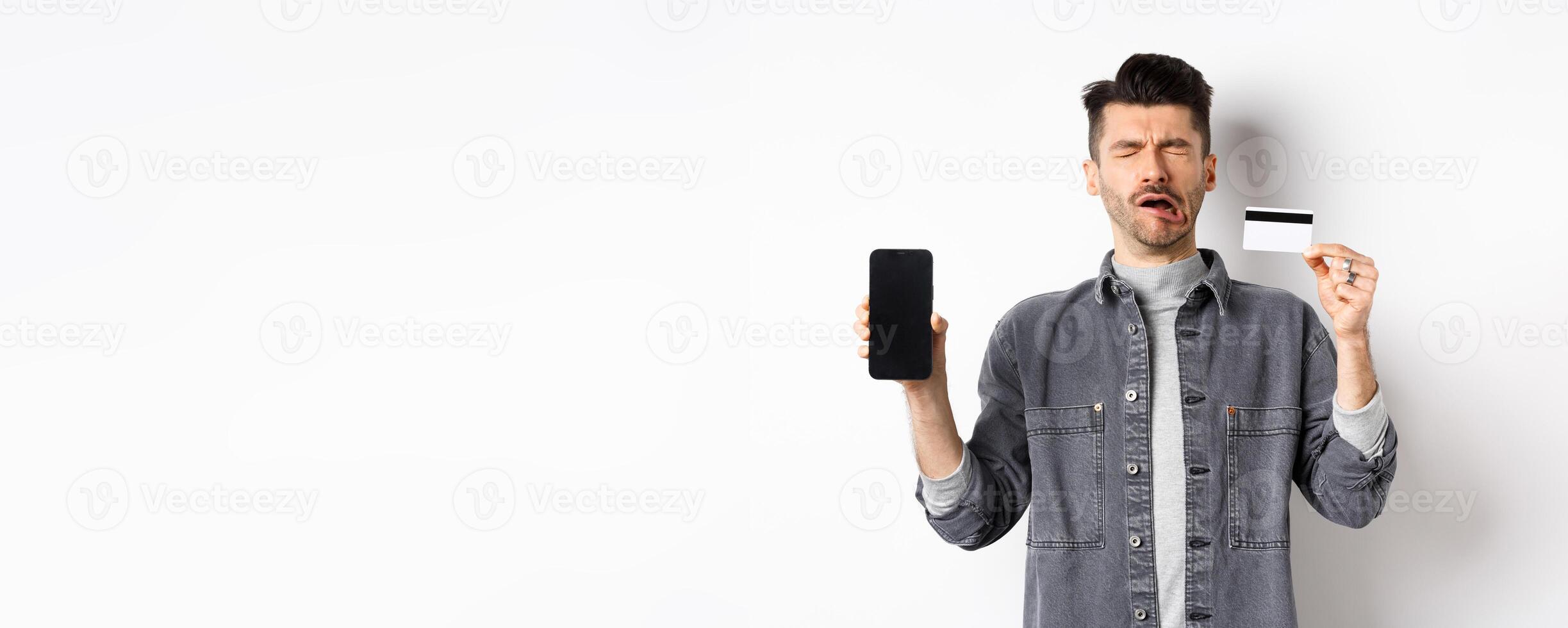 Sad crying man show empty mobile phone screen and plastic credit card, standing miserable on white background photo