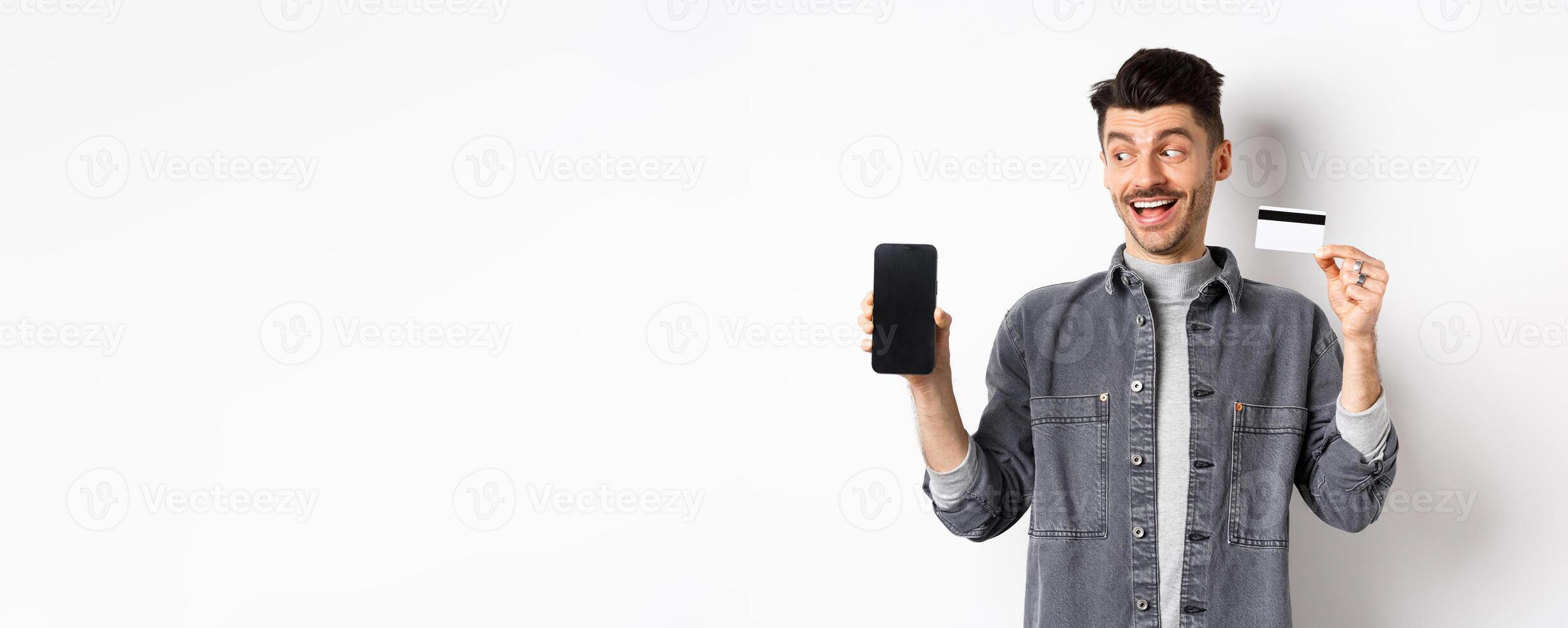 Handsome smiling man in denim jacket looking at empty smartphone screen and showing plastic credit card, good internet offer, standing against white background photo