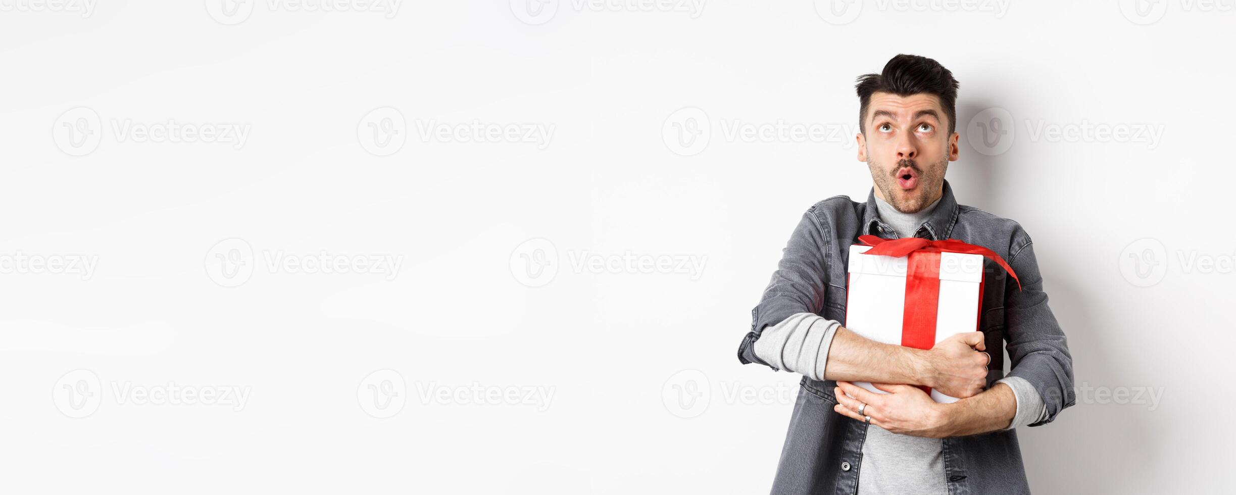 Surprised funny guy hugging big holiday gift and looking up, say wow amazed, checking out Valentines day special offer, standing on white background photo