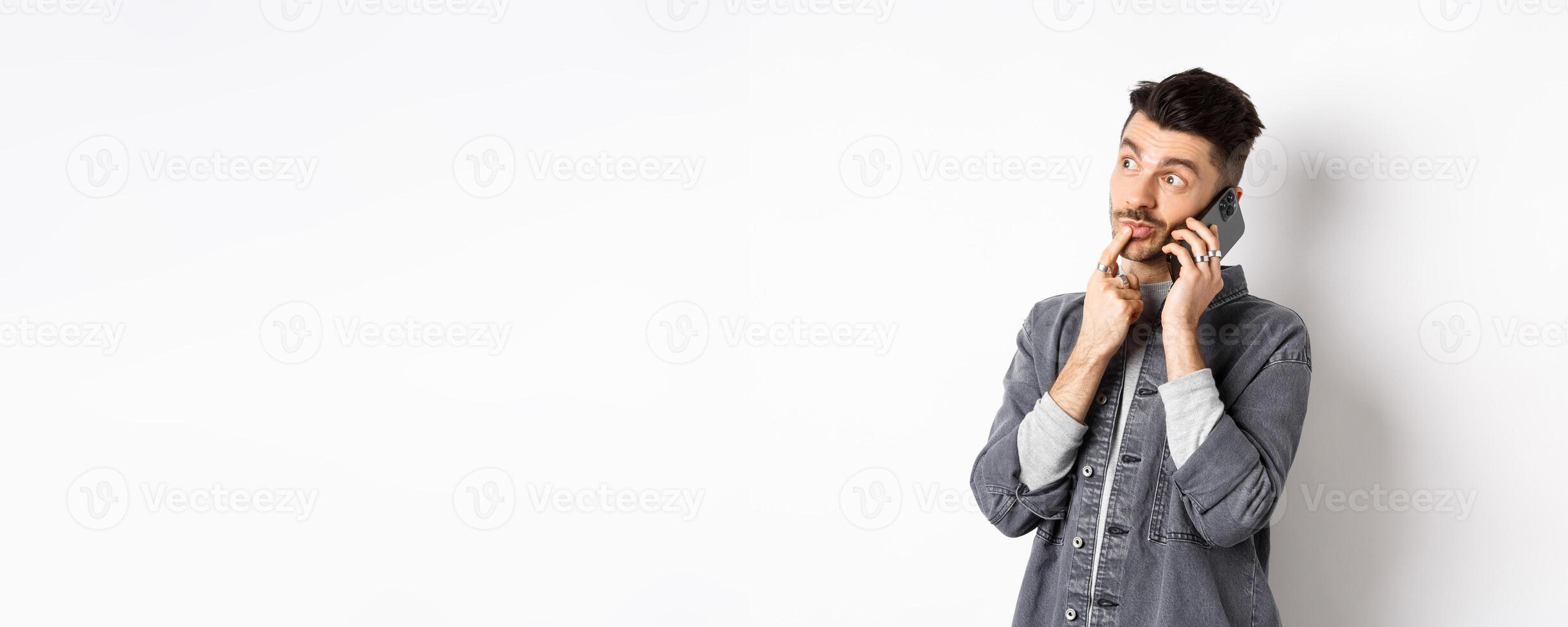Image of pensive handsome man talking on phone and thinking, making choice while making takeout order, standing against white background photo