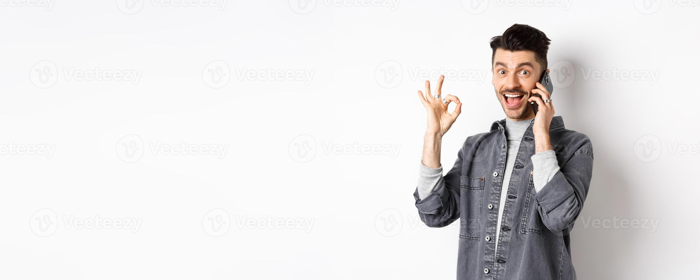 Excited young man talking on phone and showing okay gesture, smiling satisfied, standing against white background photo