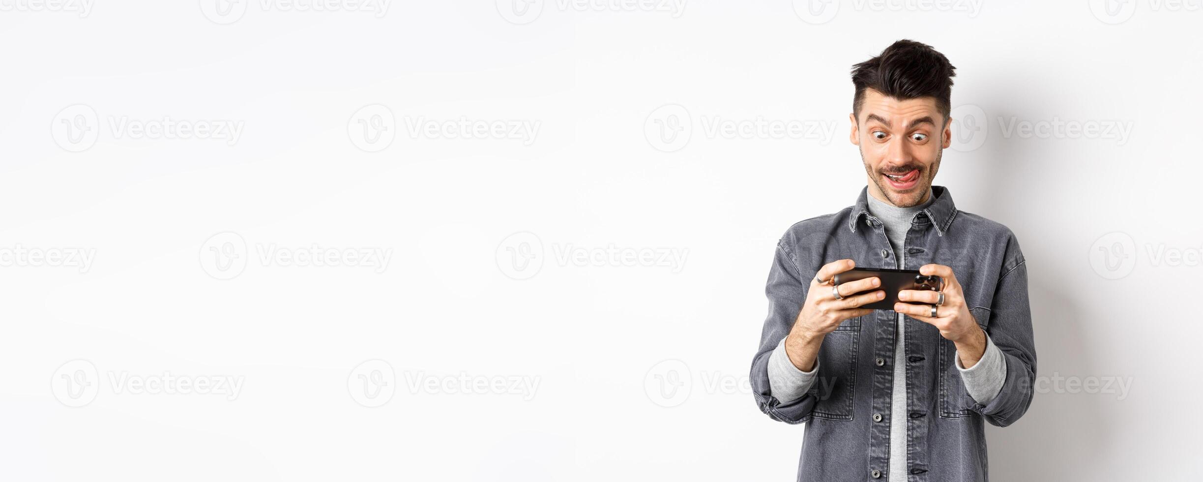 Excited guy playing game on mobile phone with focused face, tap on smartphone screen, holding cellphone horizontally, white background photo