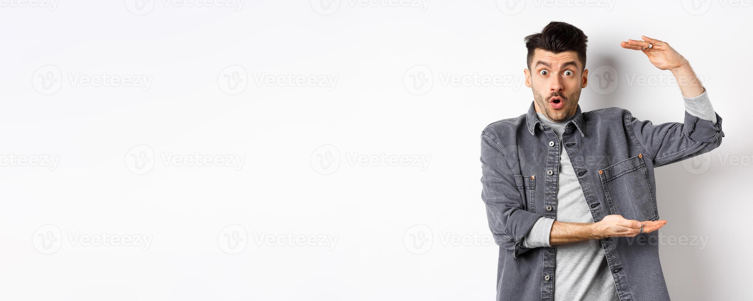 Cheerful guy smiling and showing big size, showing large thing and looking excited, standing against white background photo