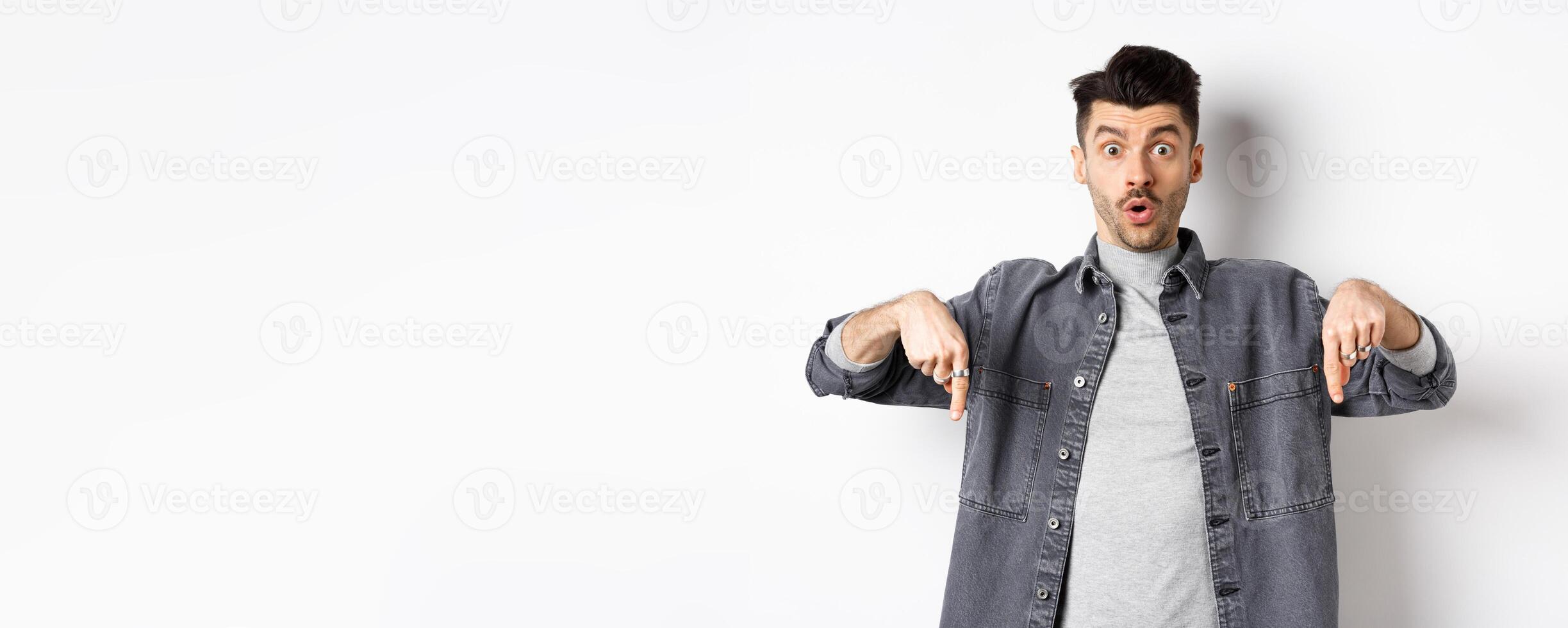 Excited guy say wow, pointing fingers down amazed, showing cool promo offer, standing on white background in denim jacket photo