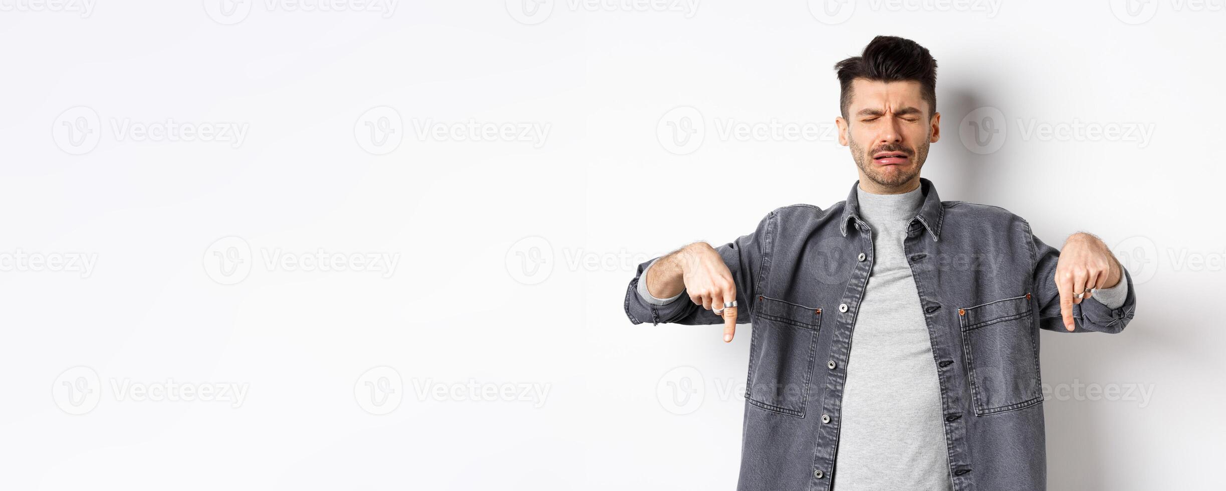Sad crying man whining, pointing fingers down disappointed, showing bad news, standing upset on white background photo