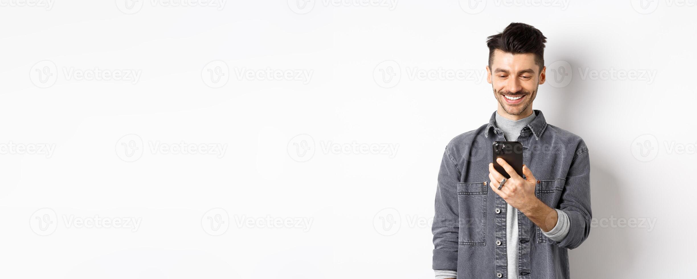 Handsome man reading message on phone and smiling. Guy holding smartphone and looking at screen, standing on white background photo
