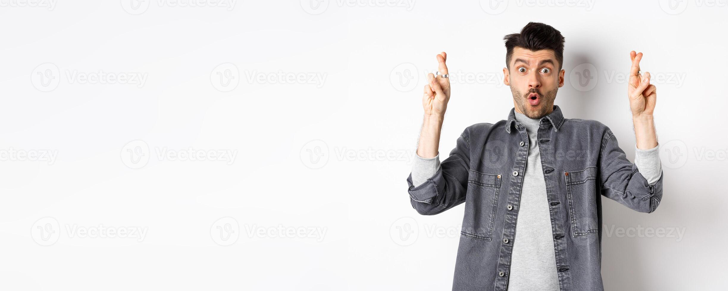 Excited young man cross fingers for good luck and gasping, holding breath as waiting important news, begging or praying, standing on white background photo