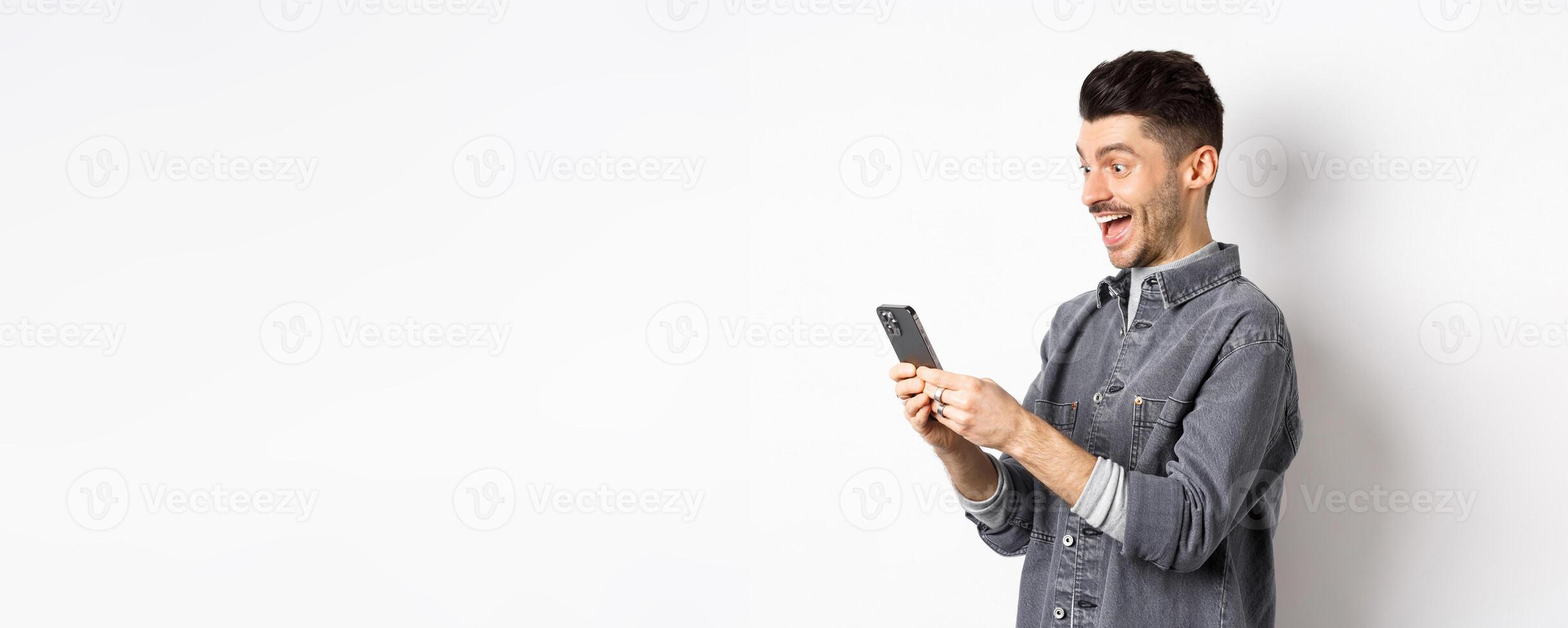 Profile portrait of excited handsome man reading message on phone, looking happy at smartphone screen, standing on white background photo