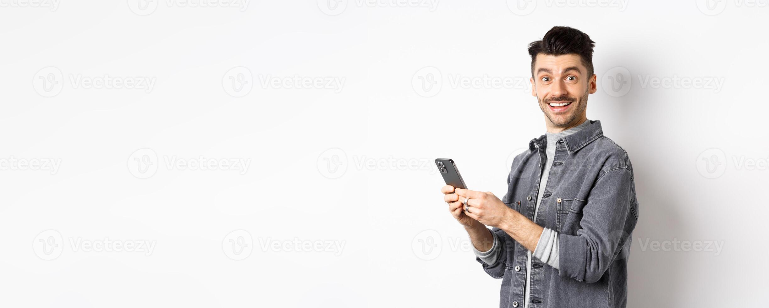 Handsome young man using smartphone and looking at camera. Caucasian guy chatting on phone with happy face, standing on white background photo