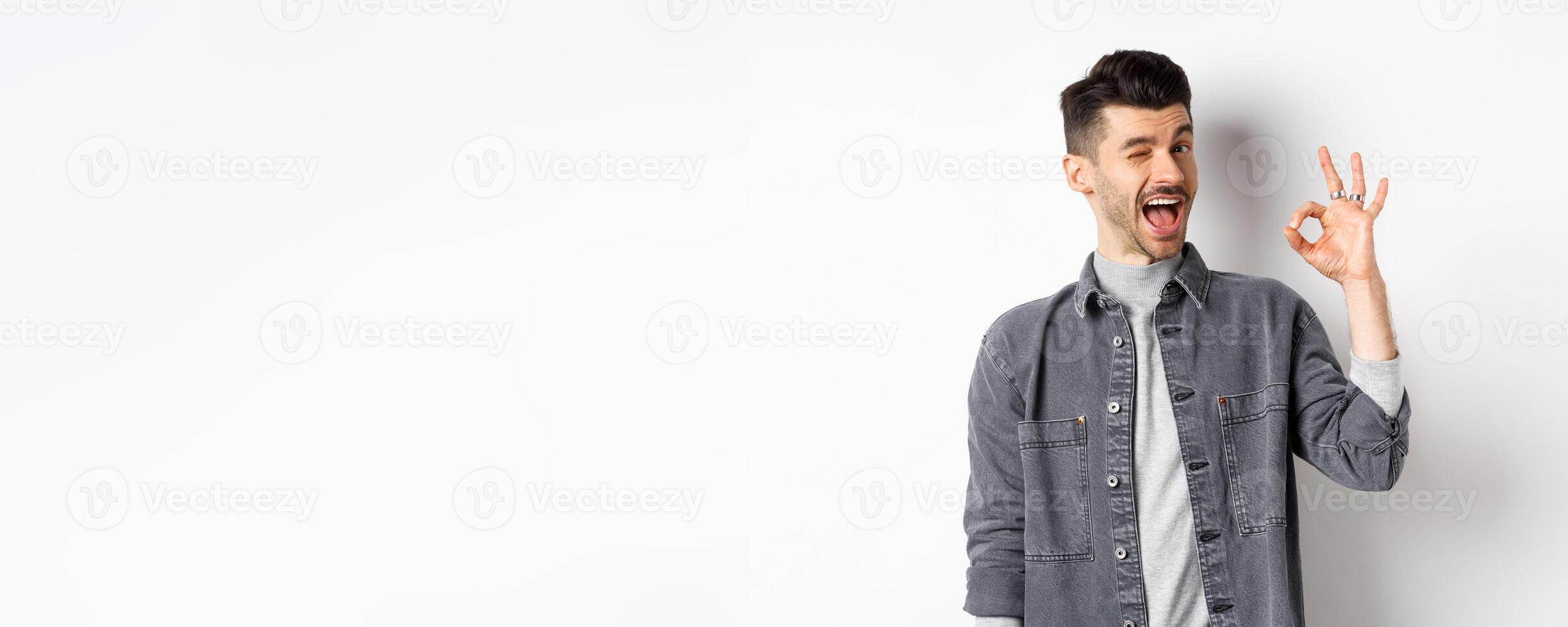 Excited handsome guy with moustache, winking and showing okay sign, smiling pleased, assure all good, praise nice work, making good job gesture, standing against white background photo