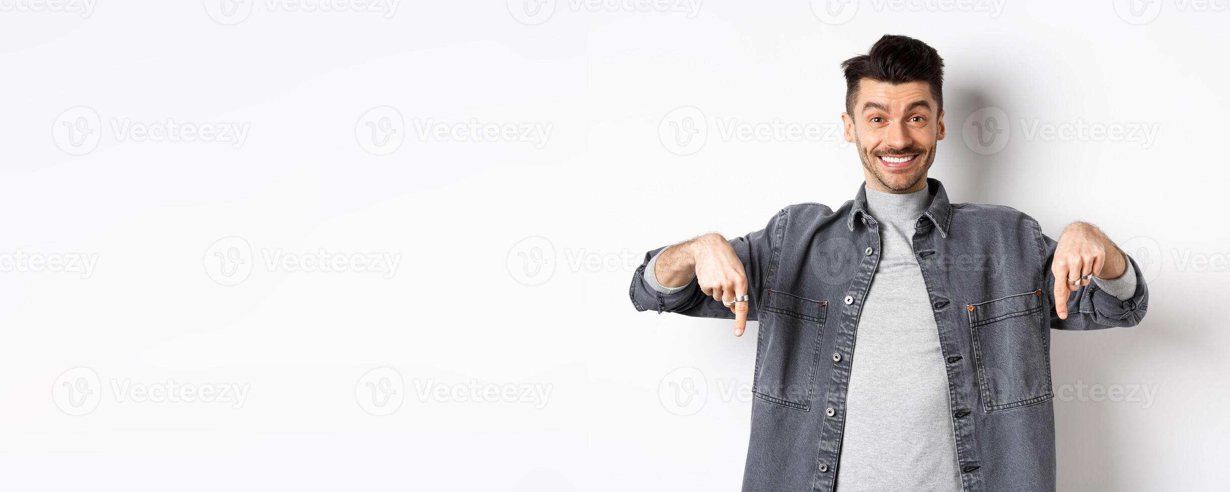 Happy caucasian man with moustache pointing fingers down, smiling and showing special deal, inviting to look here, standing on white background photo