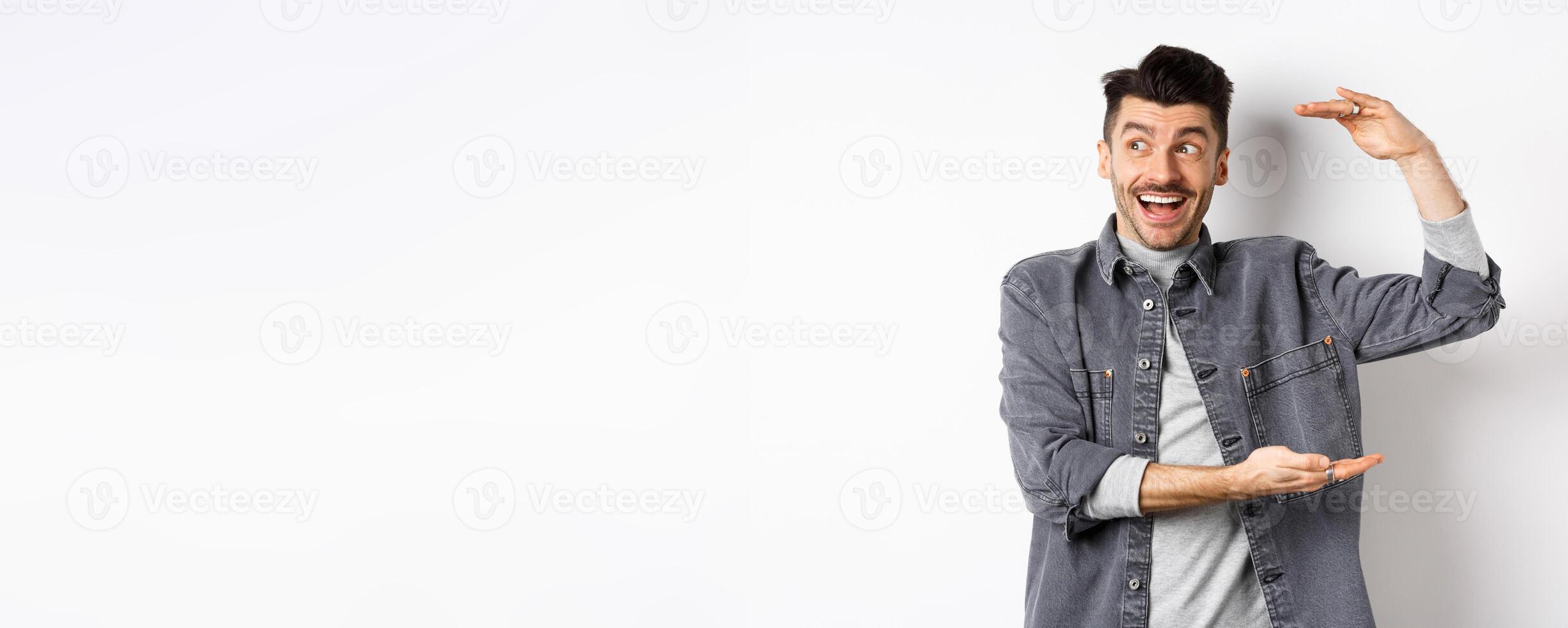 Excited guy looking at logo and showing big size thing, shaping large object and smiling amazed, standing in denim jacket on white background photo