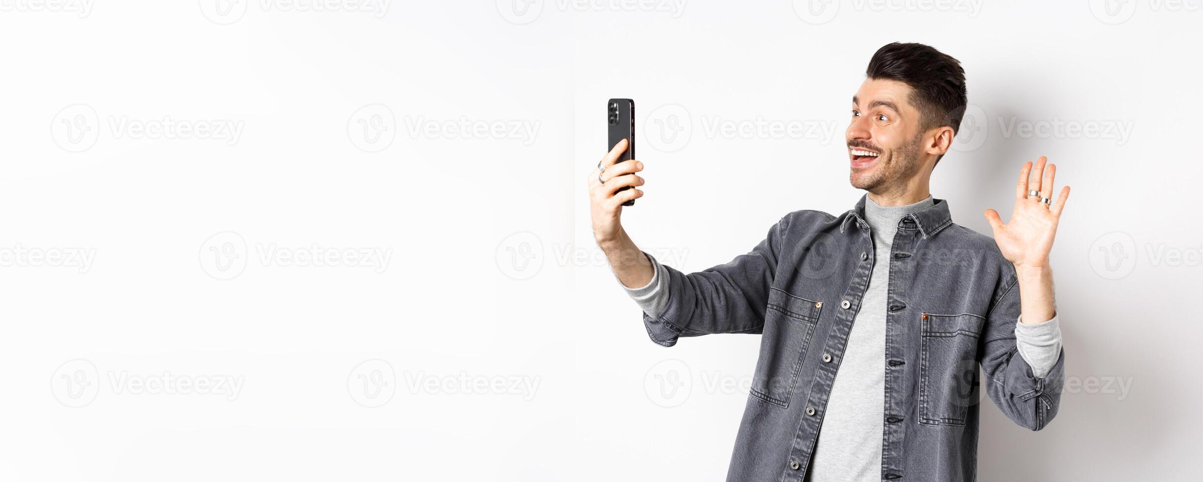Young man chatting on mobile phone, waiving hand at smartphone camera, greeting friend online, standing on white background photo