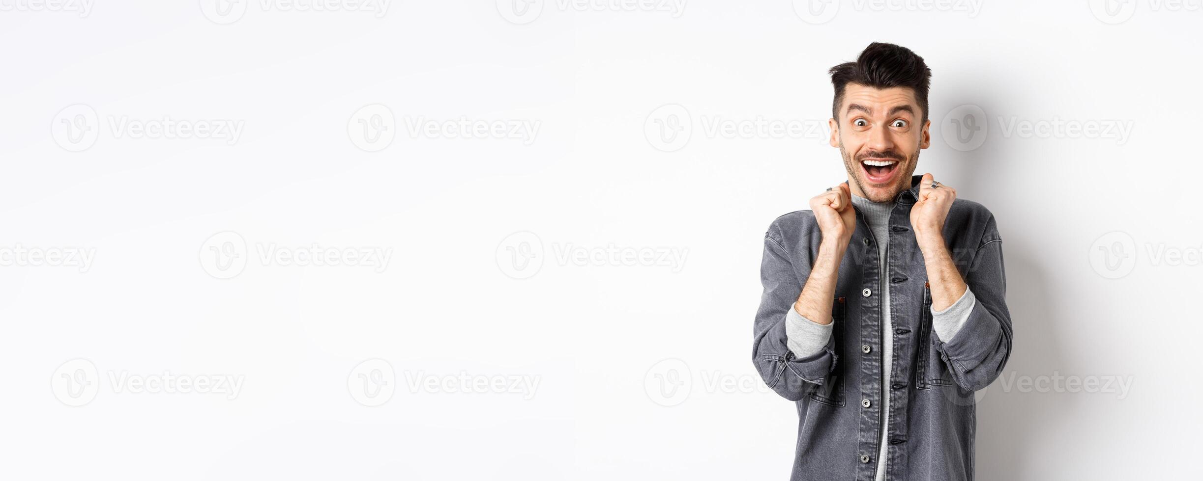 Excited young man jumping from happiness, smiling amazed and holding hands pressed to chest, standing on white background photo