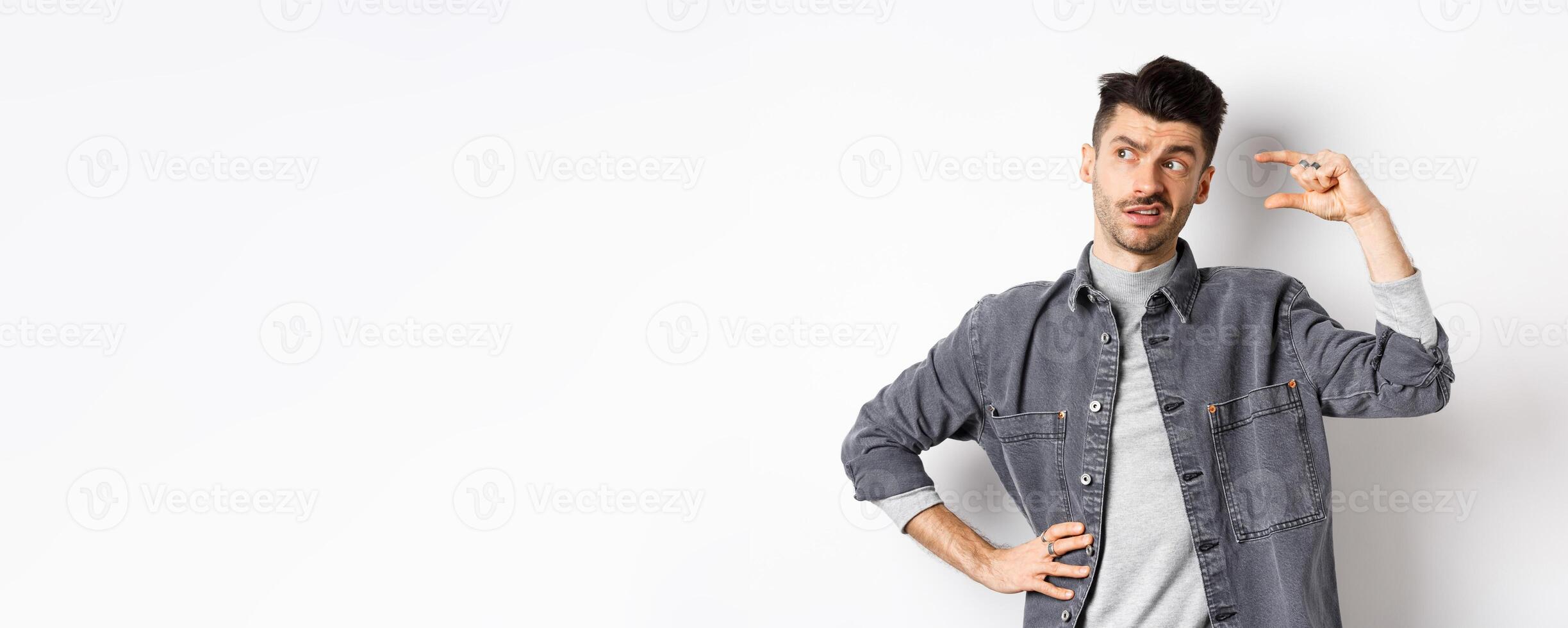Pensive young man looking aside and thinking, showing small size object, pondering something little or tiny, standing on white background photo