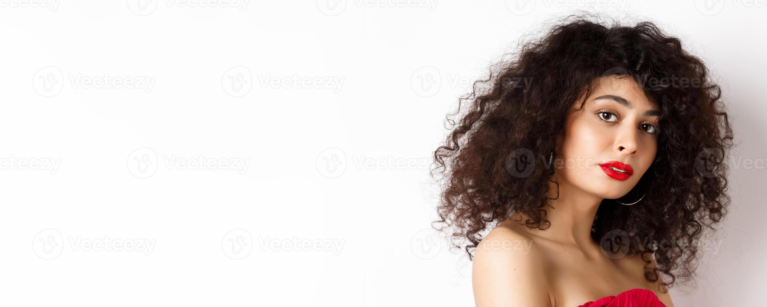 Close-up of beautiful caucasian woman with curly haircut and red lipstick, looking tender at camera, standing over white background photo
