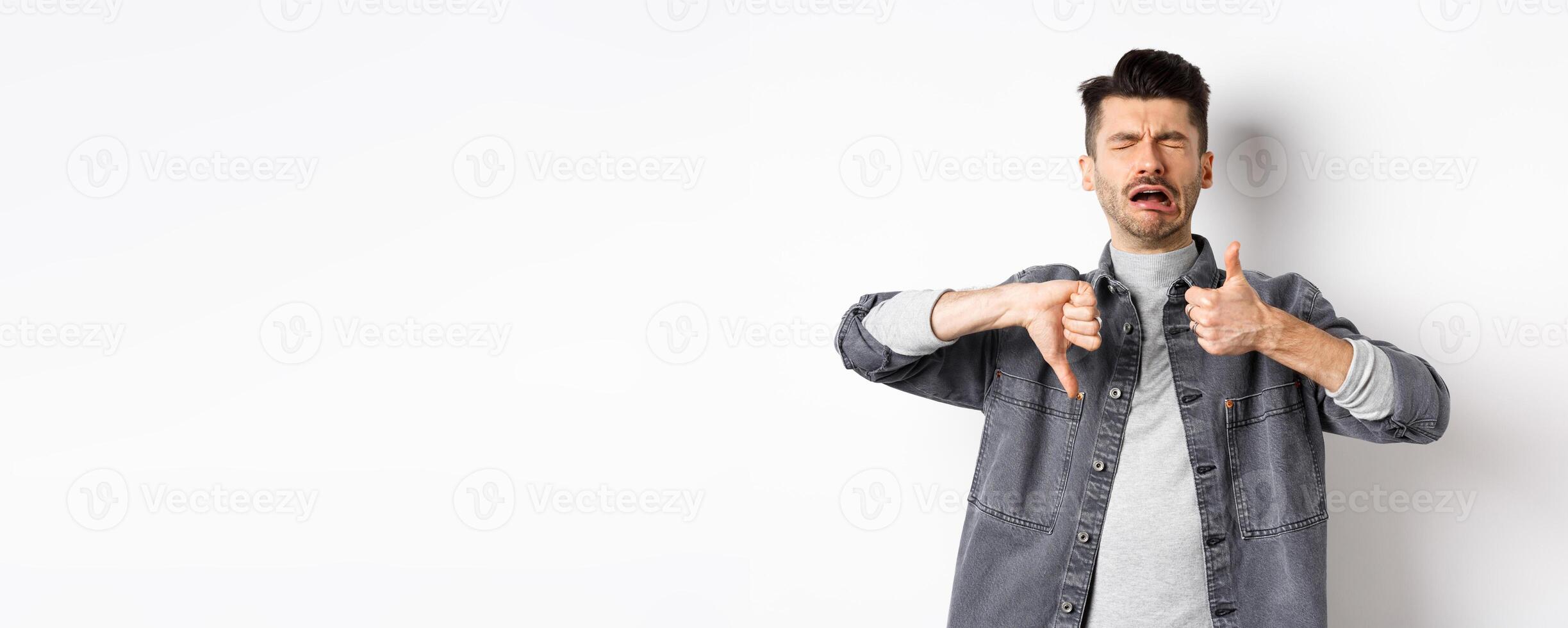 Sad man crying and showing thumbs up down, whining and complaining, standing disappointed against white background photo