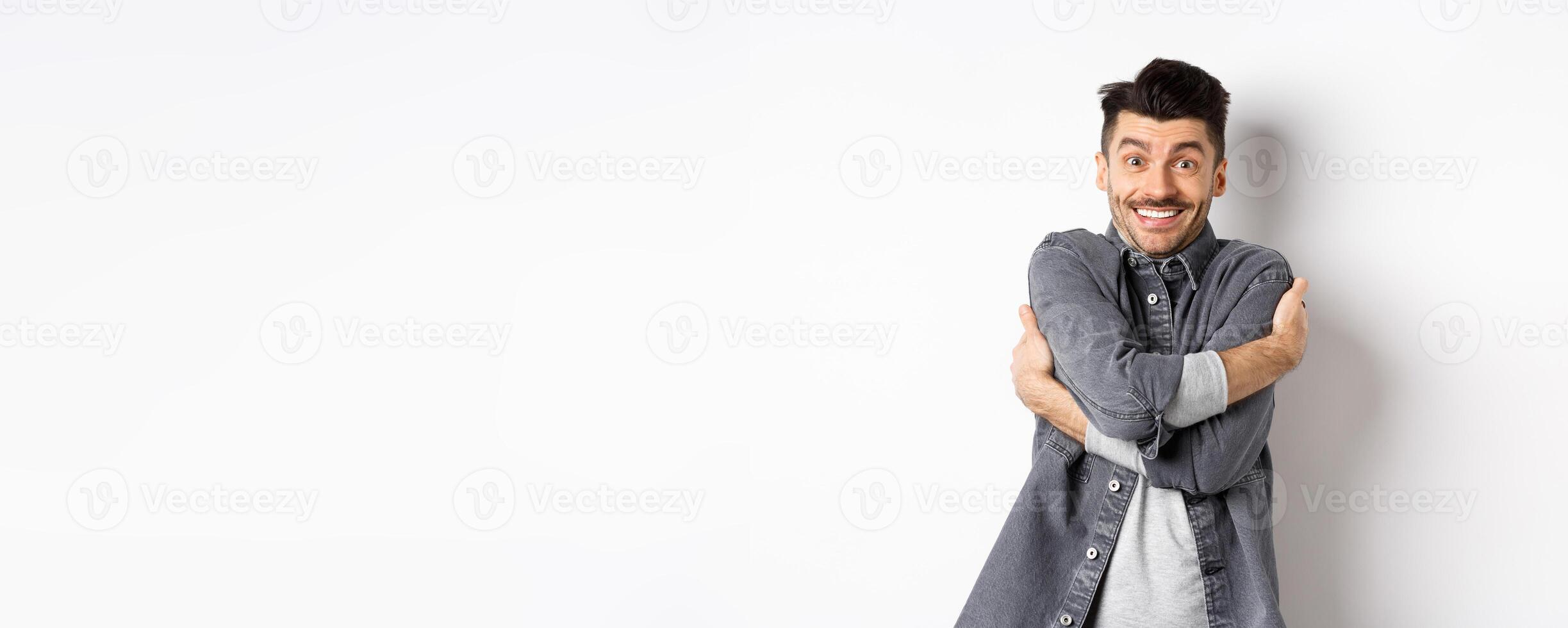 Happy handsome man hugging himself and smiling, embracing own body, standing on white background in grey jacket photo