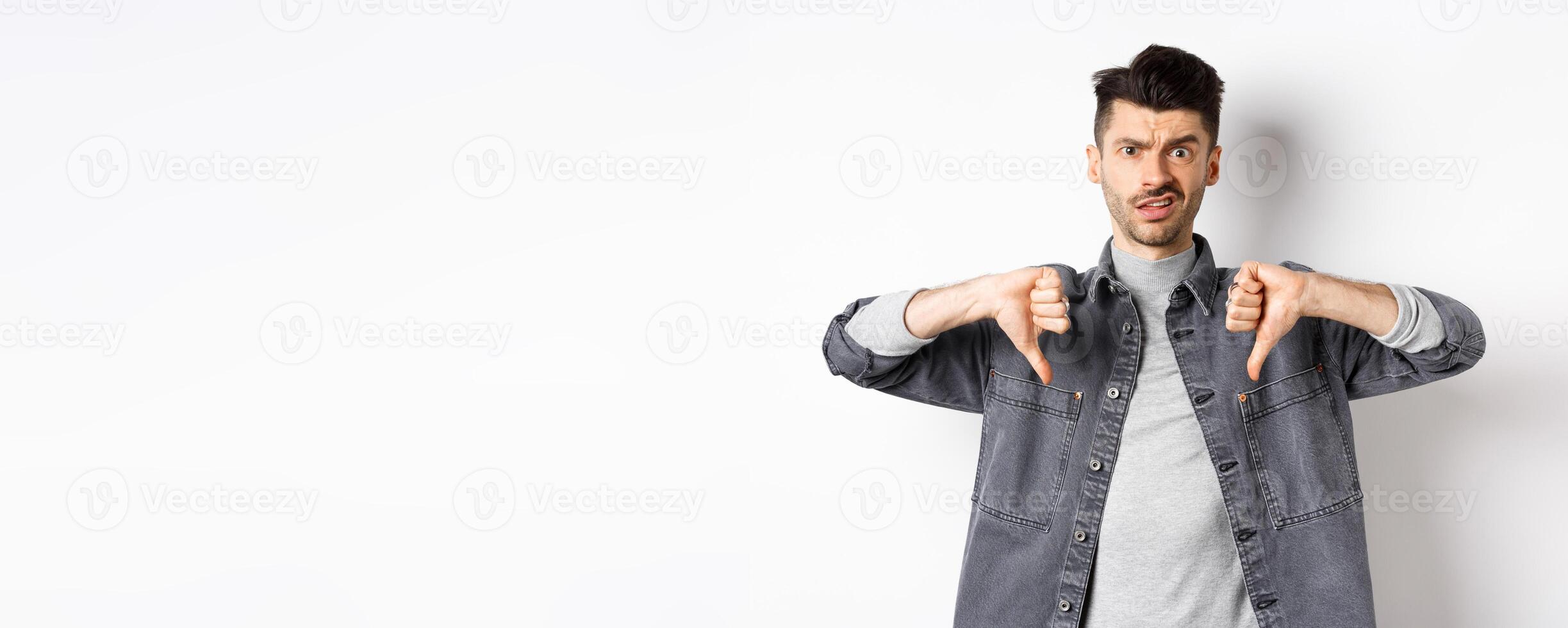 Disappointed guy showing thumbs down and frowning, looking skeptical at something bad, dislike and disapprove awful thing, standing on white background photo
