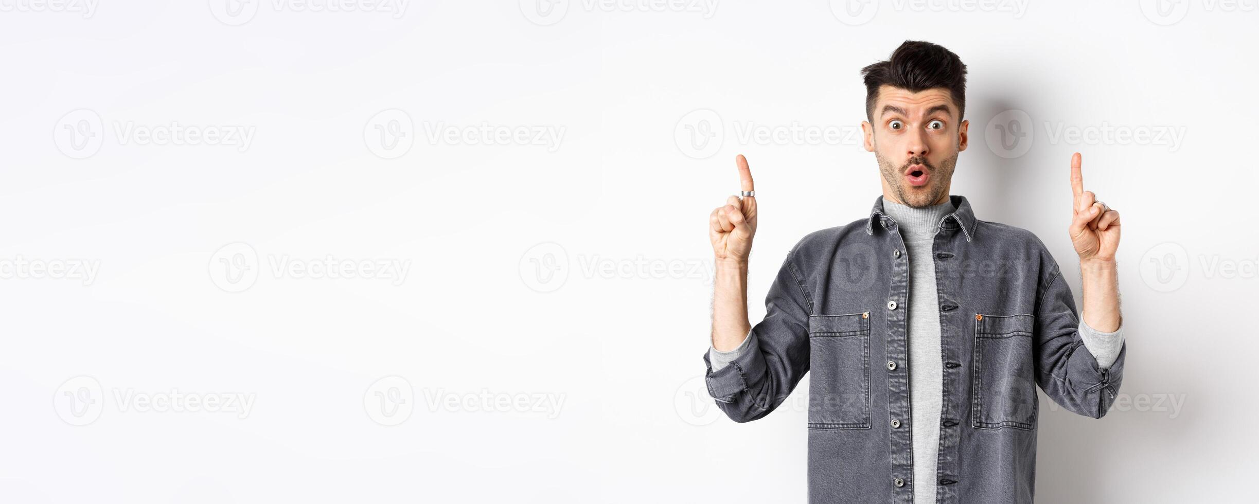 Excited man checking out promo offer, gasping amazed and pointing fingers up, making cool announcement, standing on white background photo
