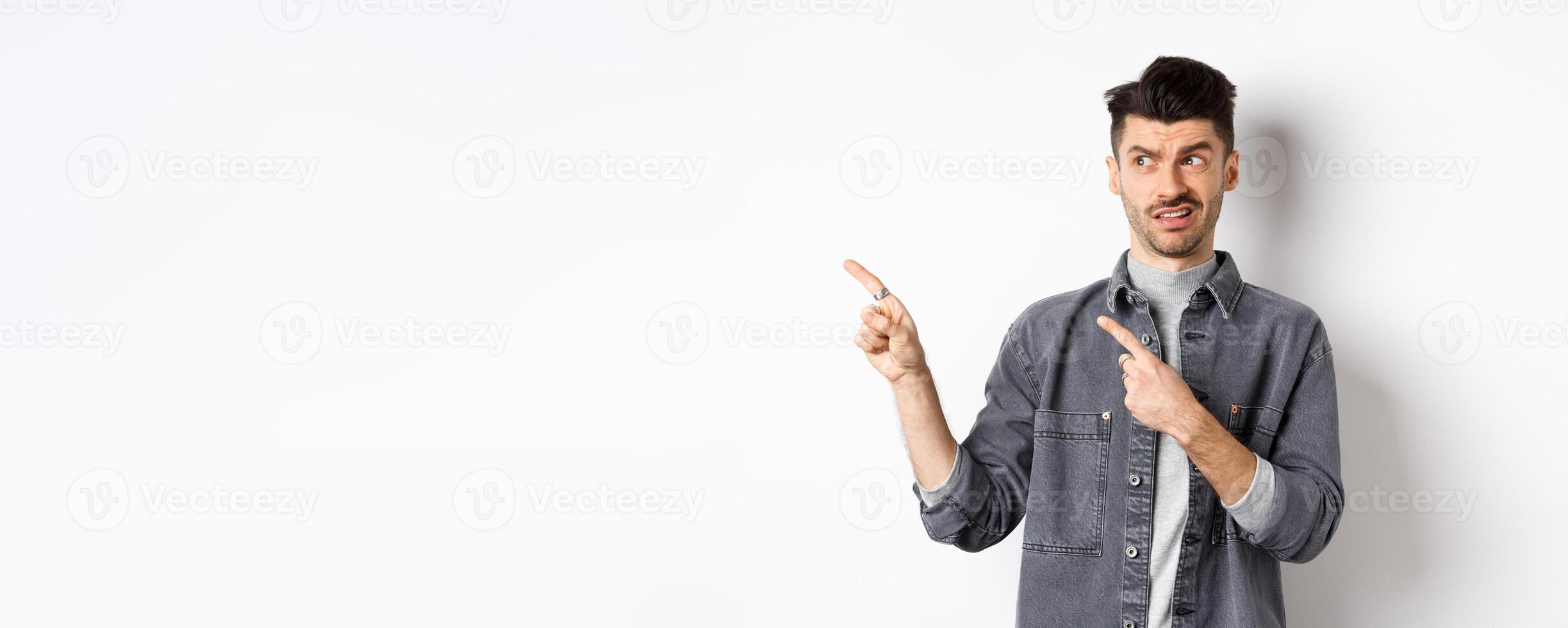Skeptical and doubtful young man pointing fingers left at logo, look with dislike and confusion, standing on white background photo