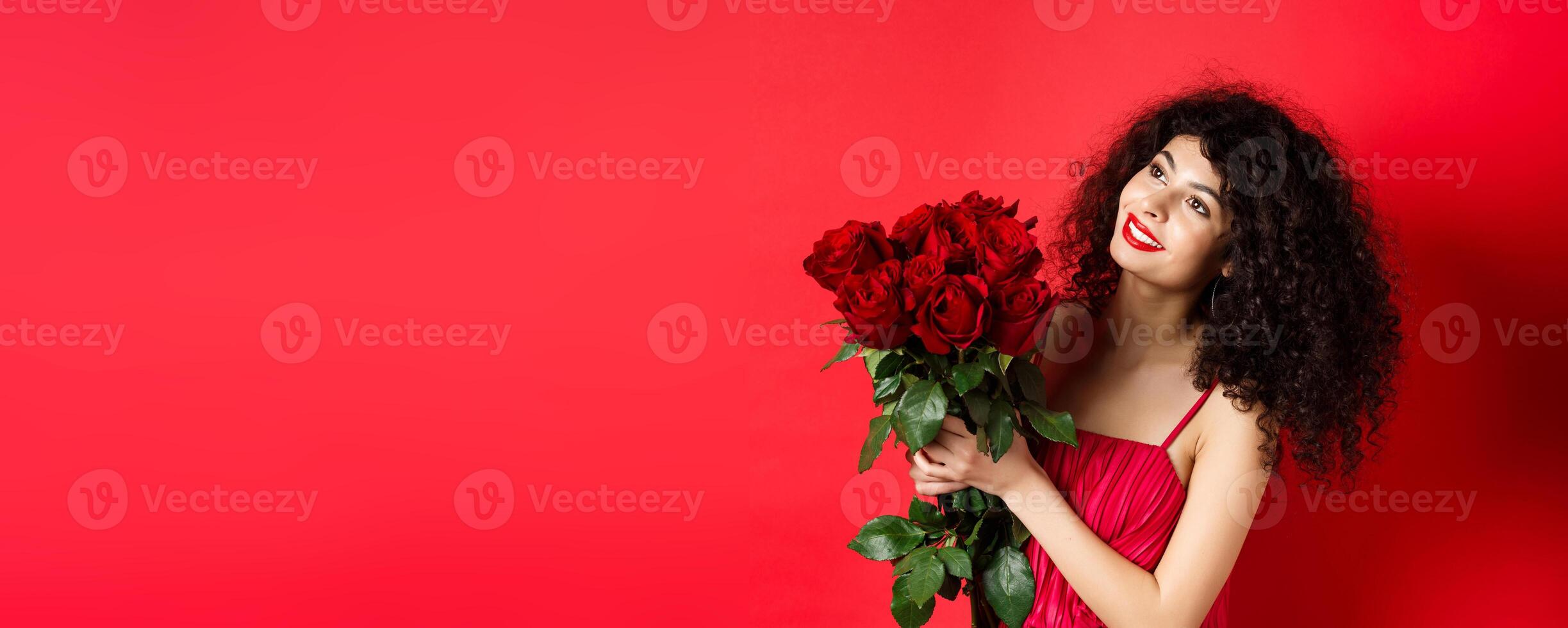 Happy beautiful woman in dress, holding flowers and smiling romantic, looking aside at logo, standing against red background photo