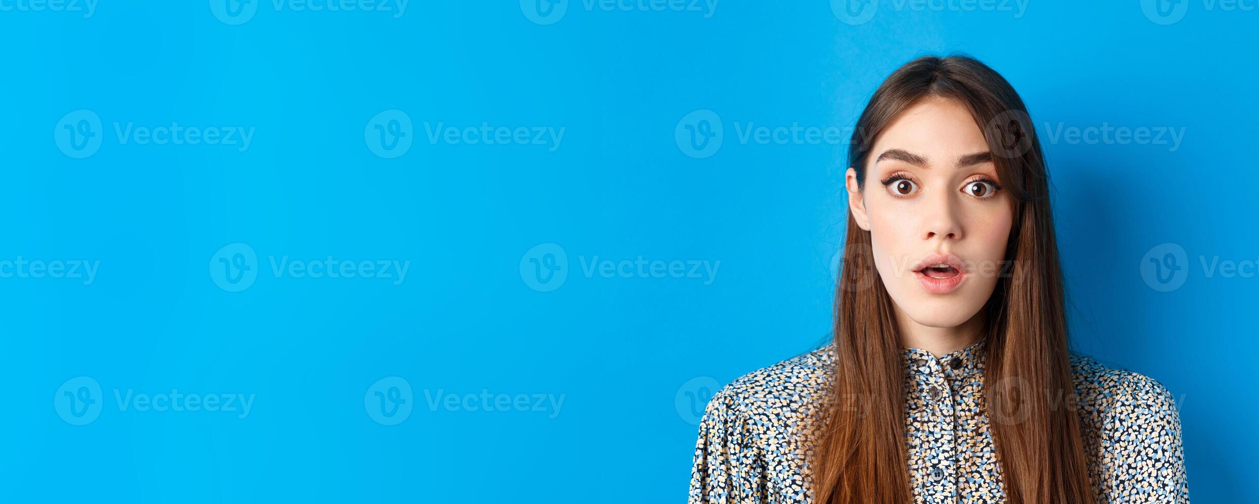 Close-up of excited pretty woman with long natural hair and makeup, drop jaw and gasping surprised, checking out awesome promo, hear amazing news, blue background photo