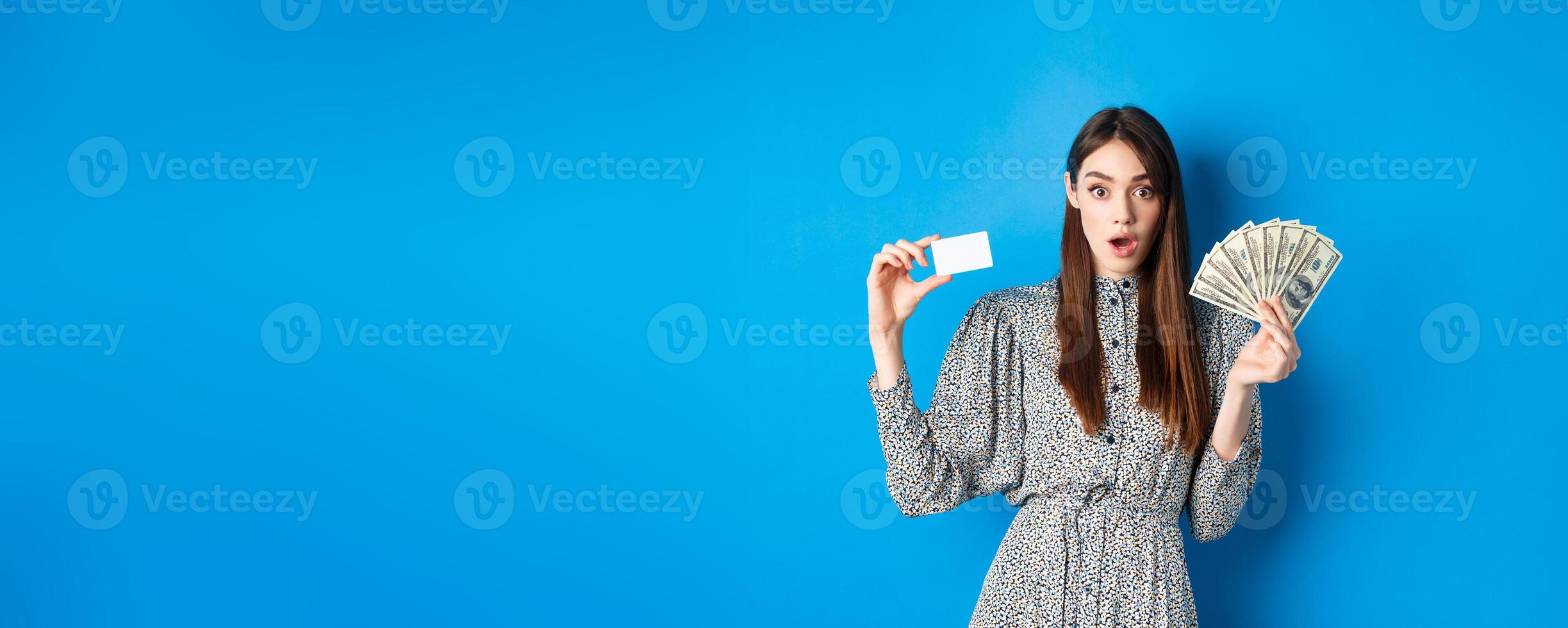 Excited lucky girl showing plastic credit card and money dollar, gasping amazed, buying something expensive, standing on blue background photo