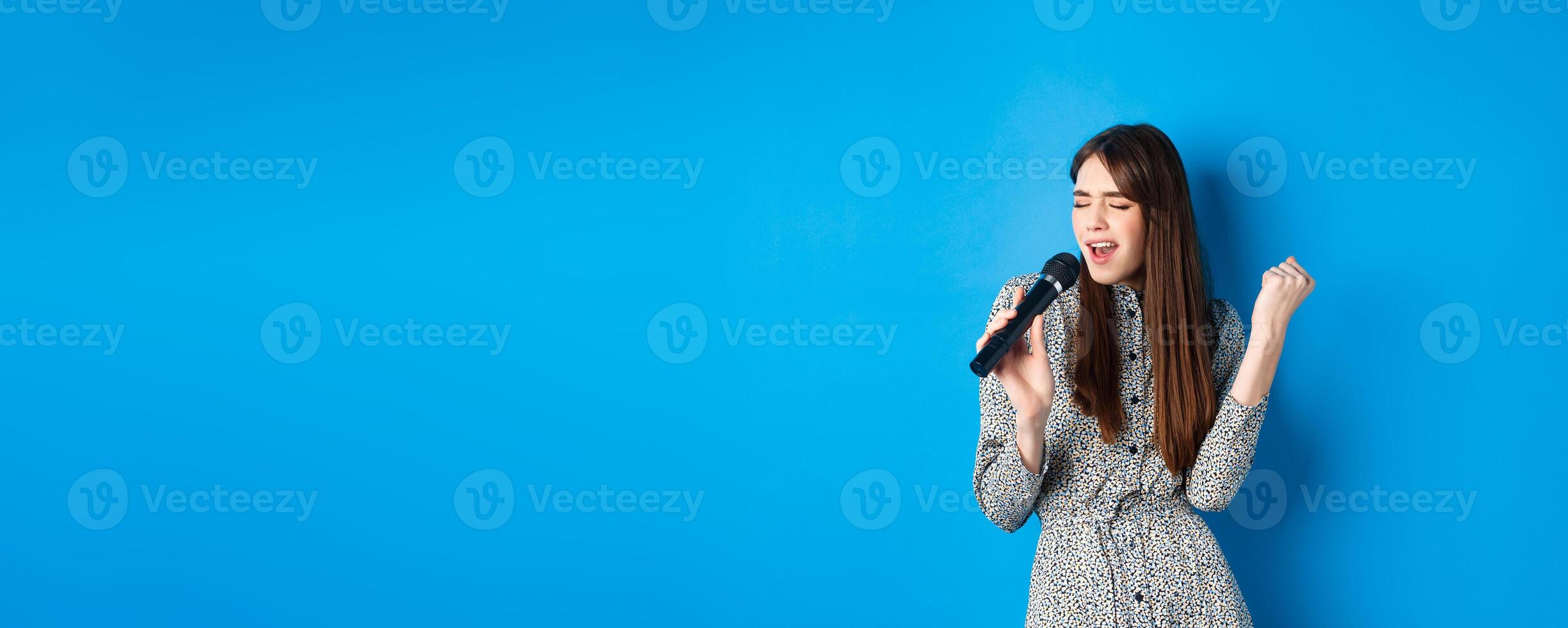 Pretty natural girl in dress singing songs in microphone, holding mic and looking passionate while performing, standing on blue background photo