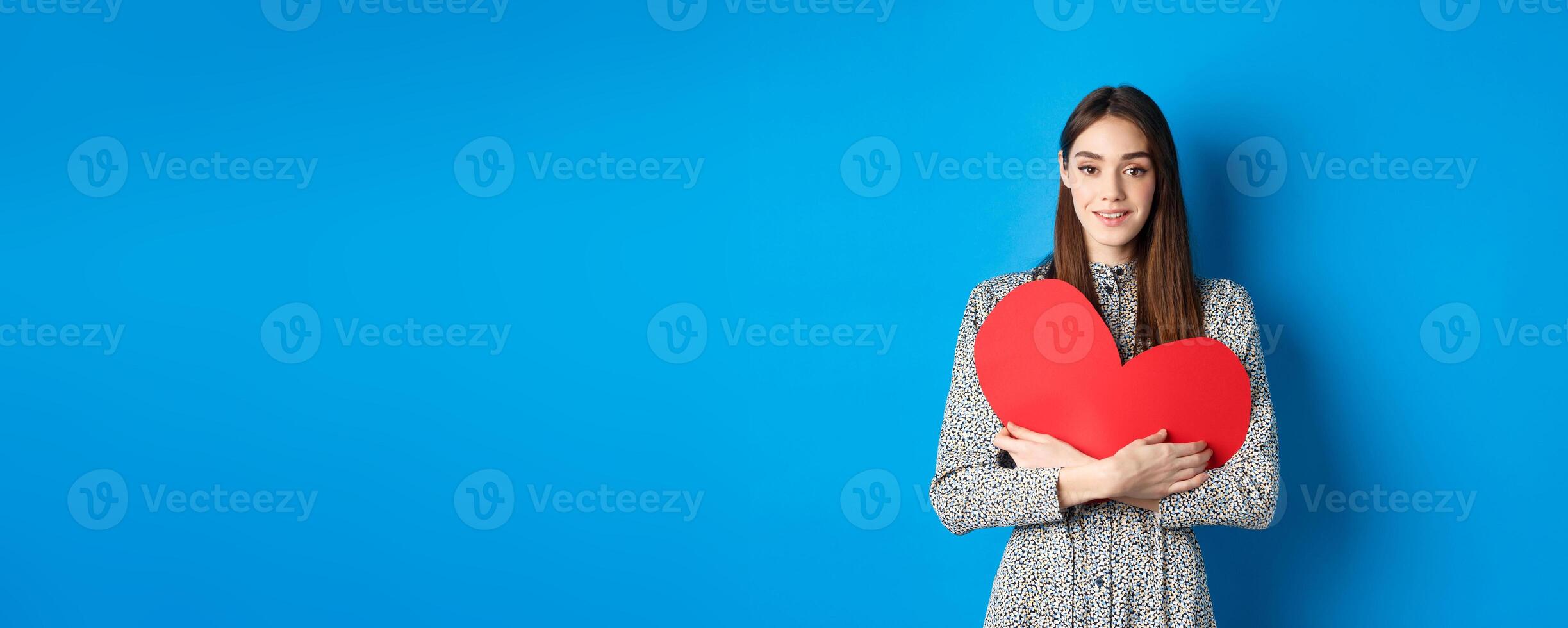Valentines day. Attractive young woman searching for love, holding big red heart cutout and smiling at camera, standing in dress on blue background photo
