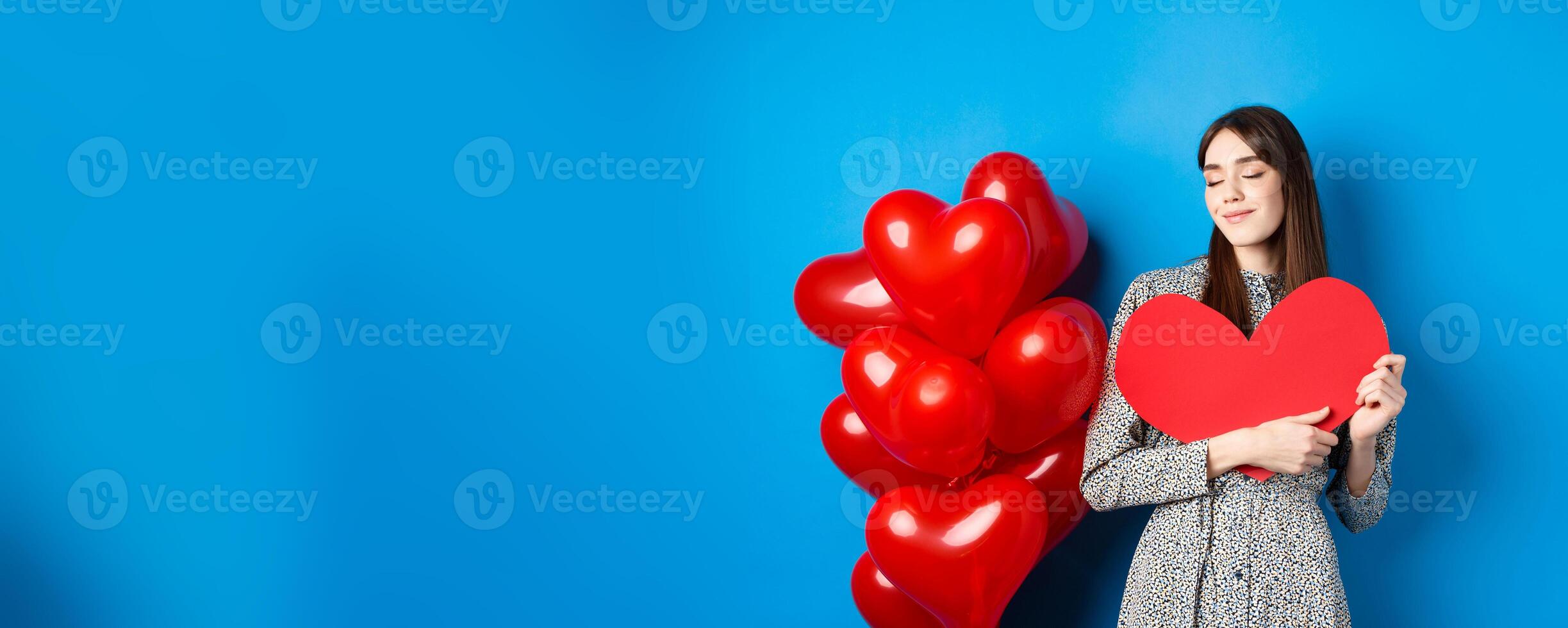 Valentines day. Romantic smiling woman hugging big red heart cutout and dreaming of love, standing in dress on blue background photo