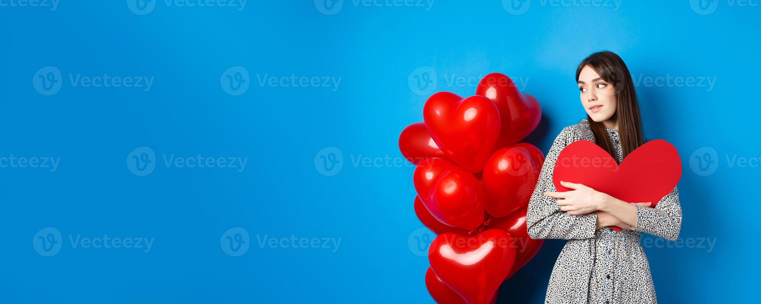 Valentines day. Beautiful and romantic woman looking pensive at balloons, hugging big red heart and smiling, waiting for love, blue background photo