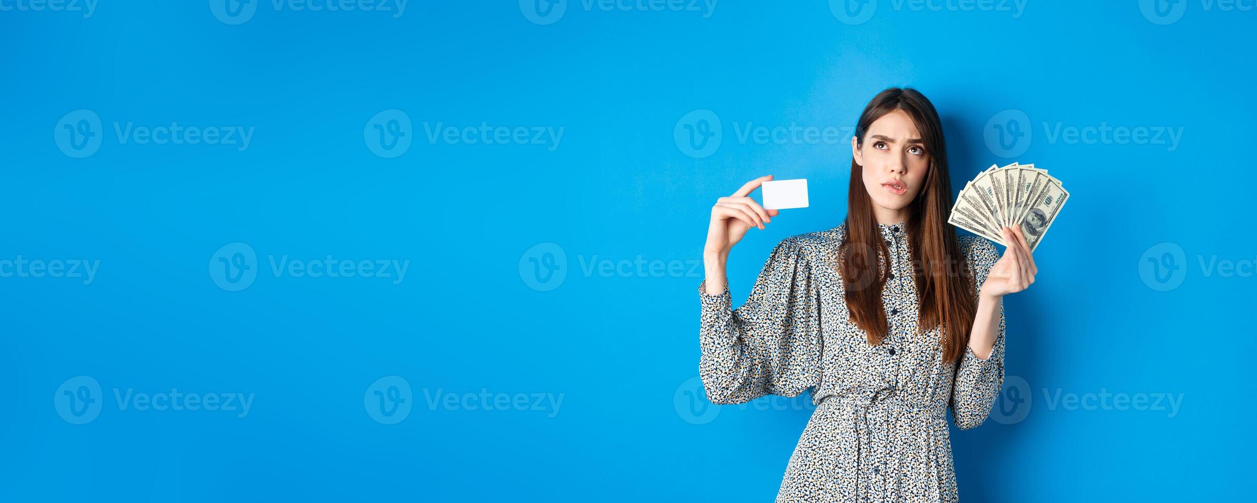 Shopping. Thoughtful girl looking up and biting lip, counting in mind or thinking, holding plastic credit card with dollar bills, standing on blue background photo