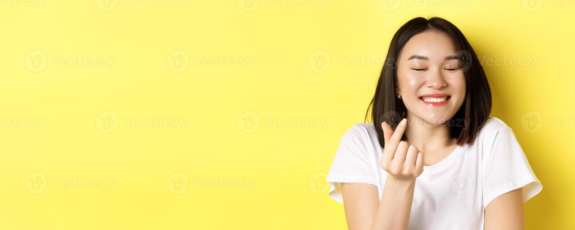 Valentines day and women concept. Close up of pretty asian girl in white t-shirt, smiling and showing finger heart, I love you gesture, standing over yellow background photo